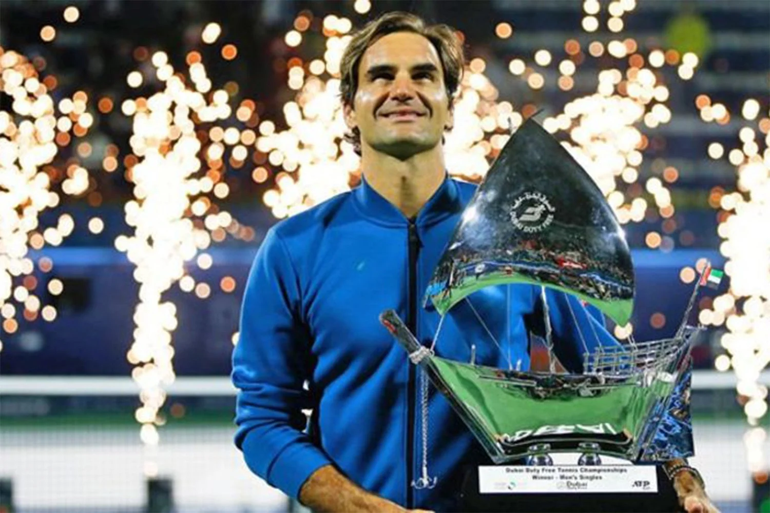 A man in a blue jacket holding a reflective trophy with fireworks in the background.