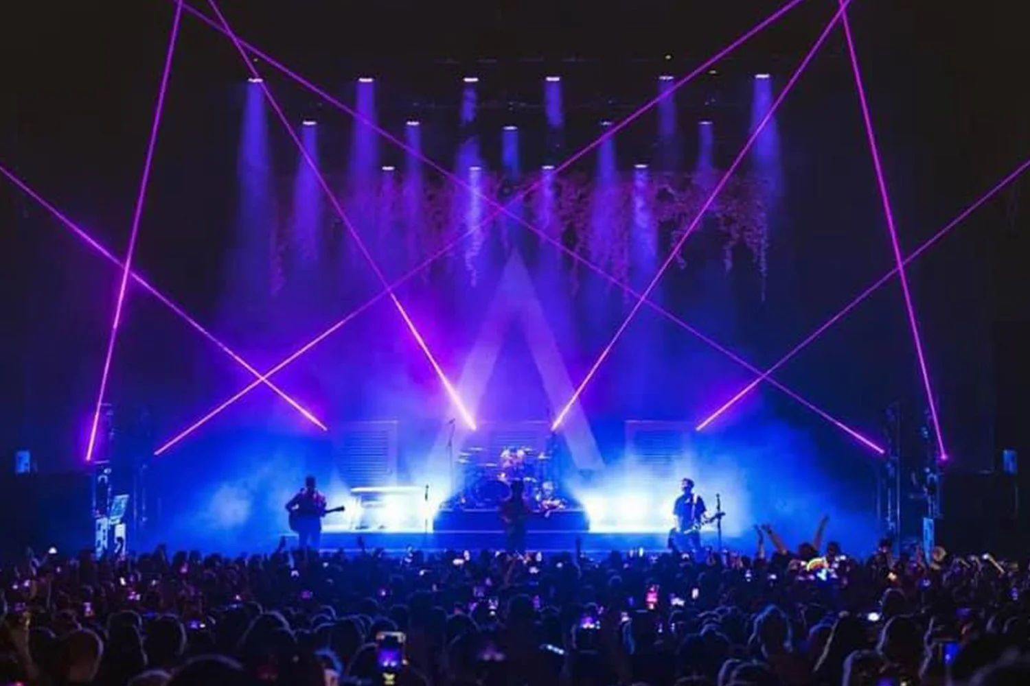A concert stage with bright purple and blue lights, laser beams, and a crowd watching the performance.