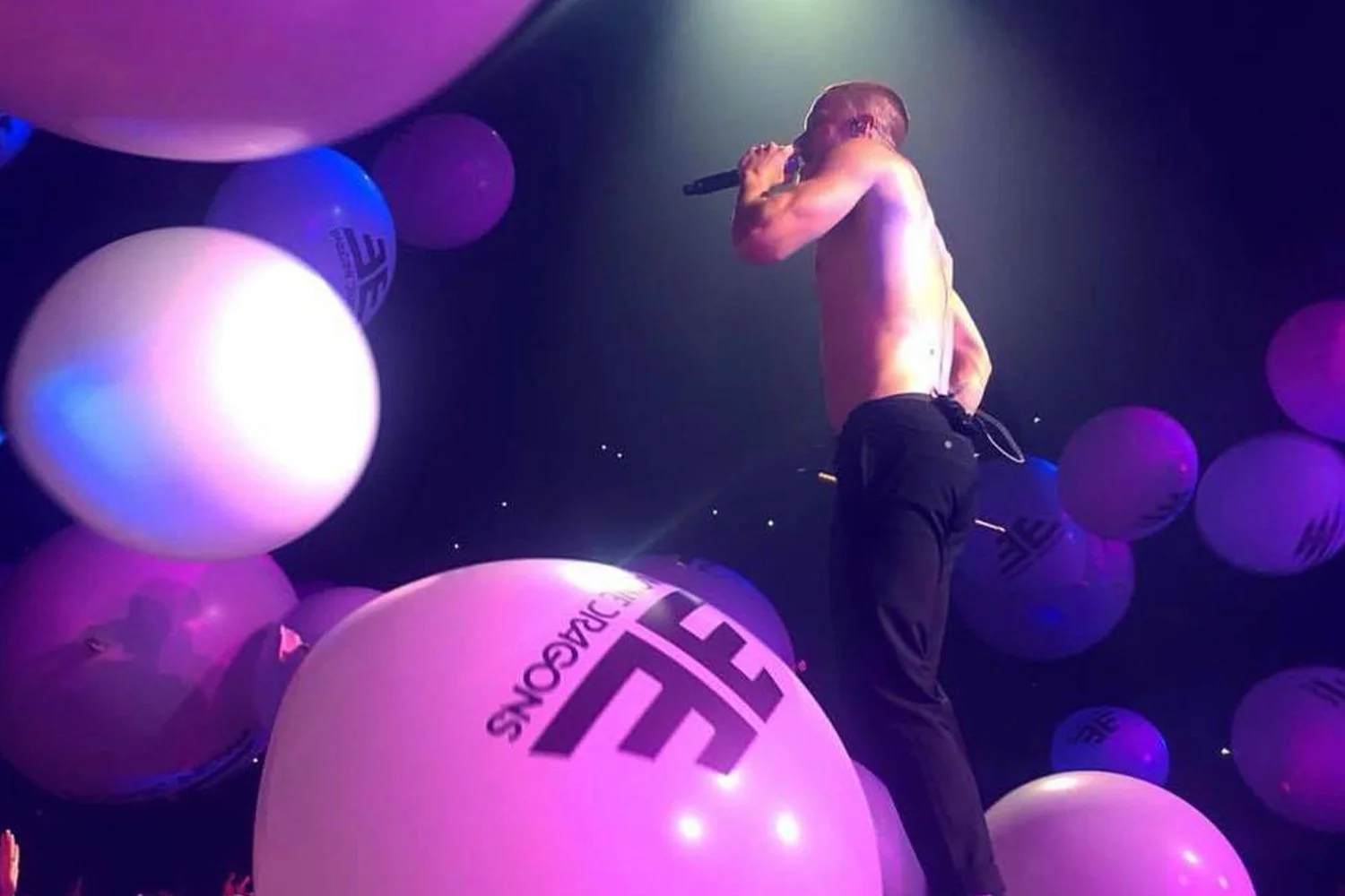 A shirtless man holding a microphone on stage surrounded by large purple and white balloons with some light shining on him in a dark environment.