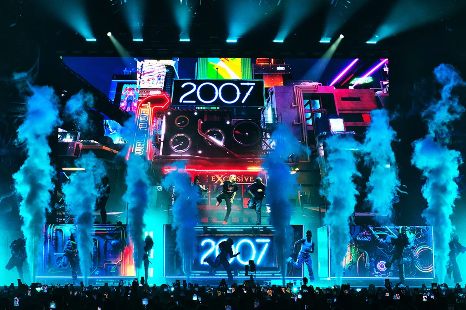 A concert stage with bright neon lights, large digital screens displaying '2007', smoke effects, and dancers performing.
