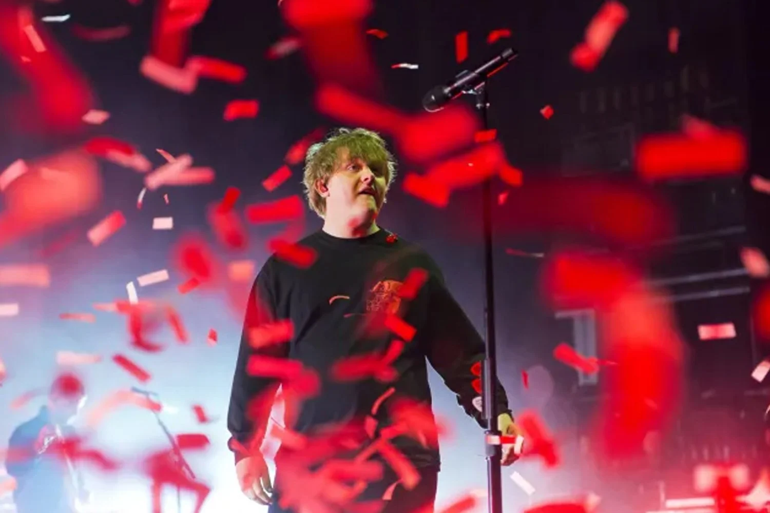 A young man with curly blonde hair standing on stage surrounded by red and white confetti, wearing a black shirt, with a microphone and backing musicians in the background under concert lighting.