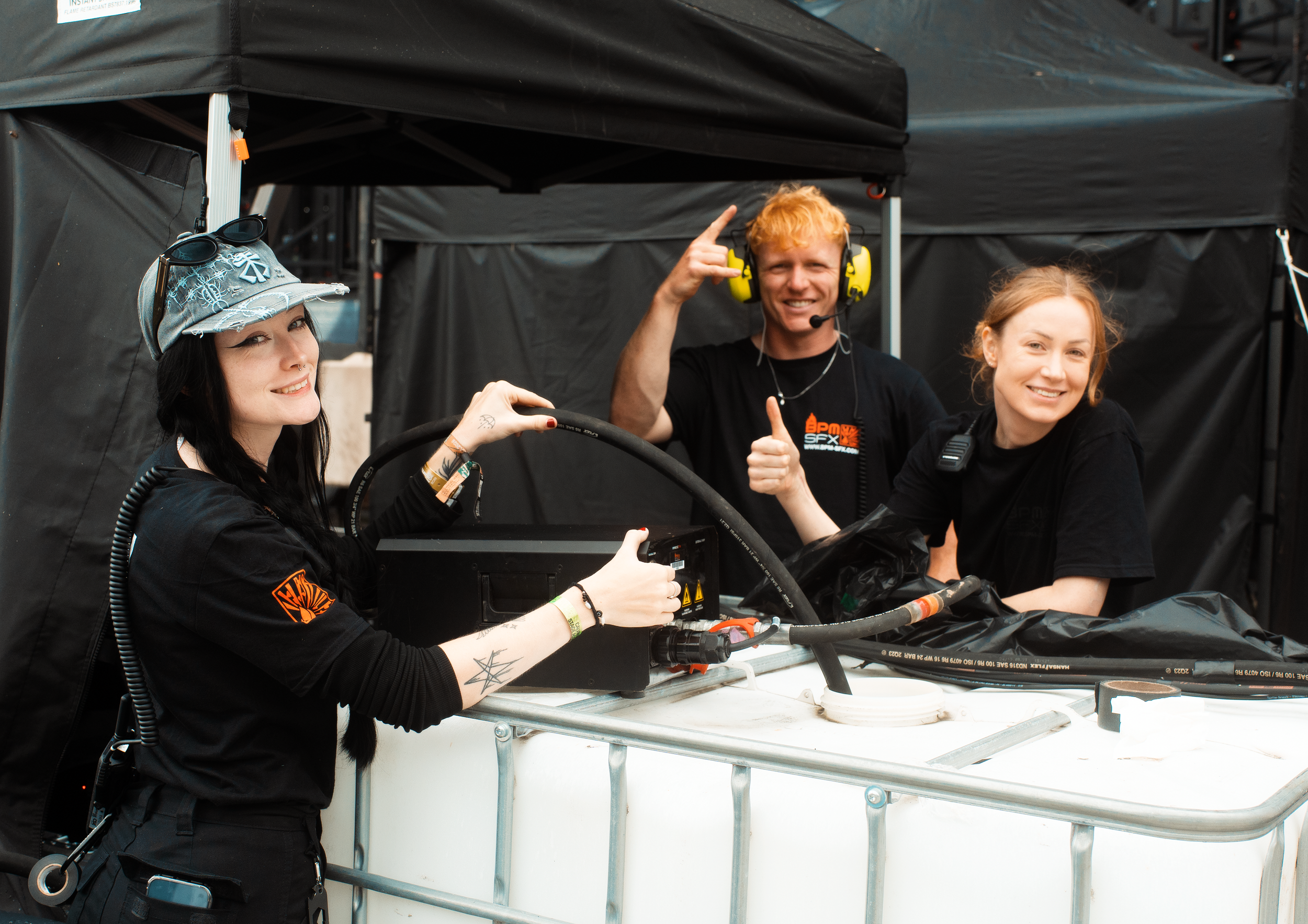 Three crew members at a race track, smiling and posing. One woman in the foreground wears a baseball cap with sunglasses, the man in the center has orange hair, is wearing headphones, and making a rock and roll hand gesture. The woman on the right is smiling and looking at the camera.