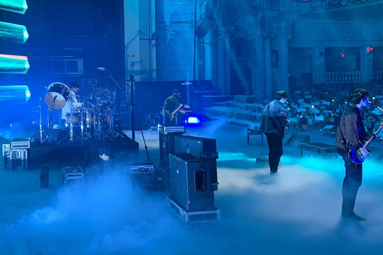 A band performing on stage with musical instruments, fog, and blue lighting, set in a venue with a seated audience.