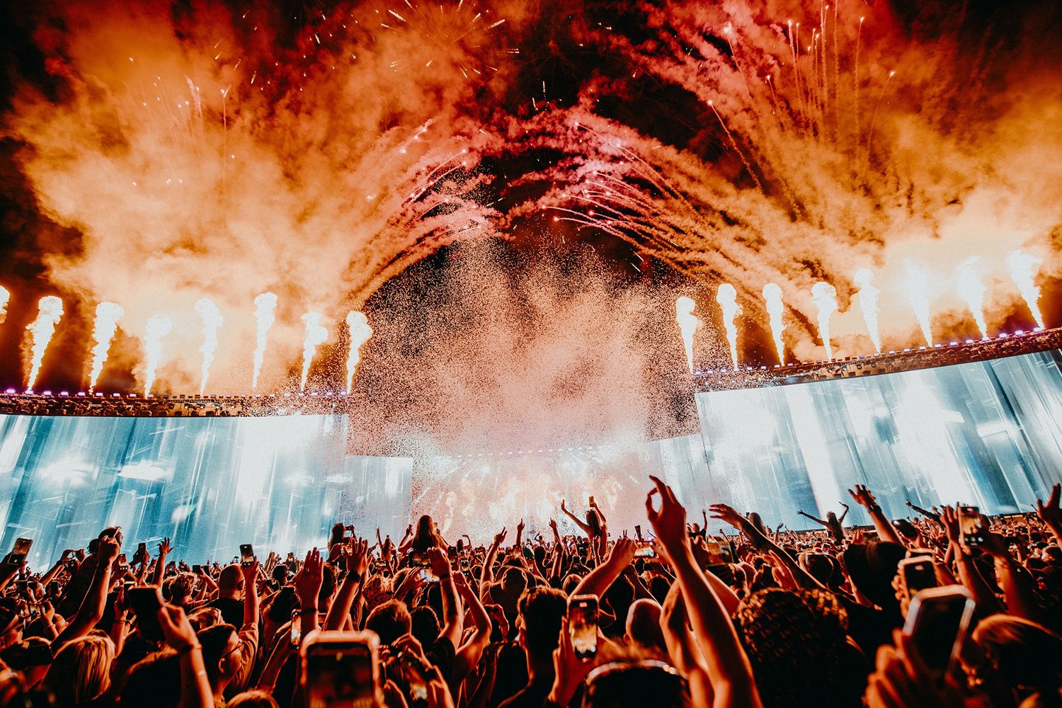 Crowd at a concert or festival with fireworks and pyrotechnics lighting up the night sky, people raising hands and taking photos.