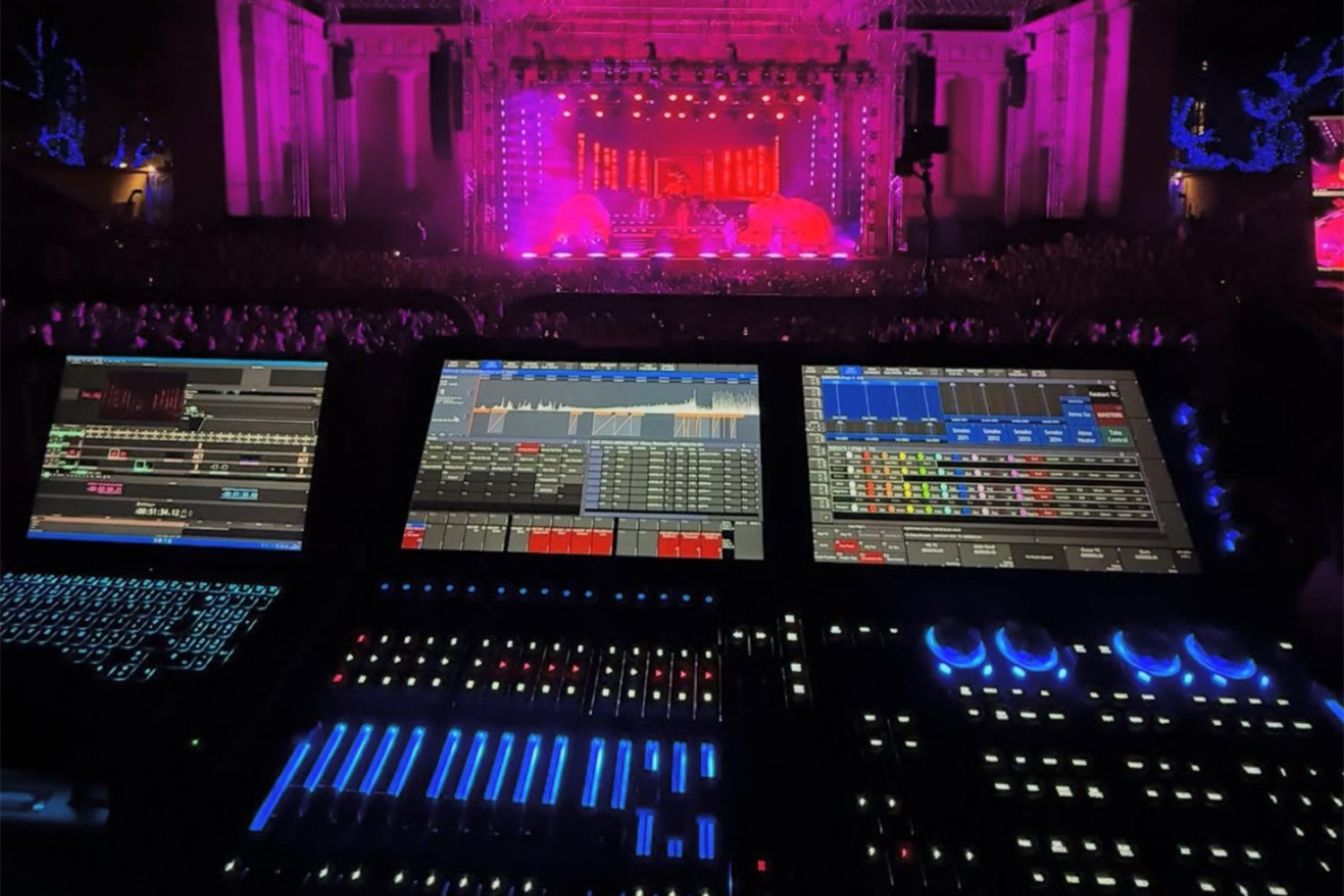 Music concert stage illuminated with pink and purple lights, large crowd in front, electronic music DJ setup with LED screens and lighting controllers in the foreground.