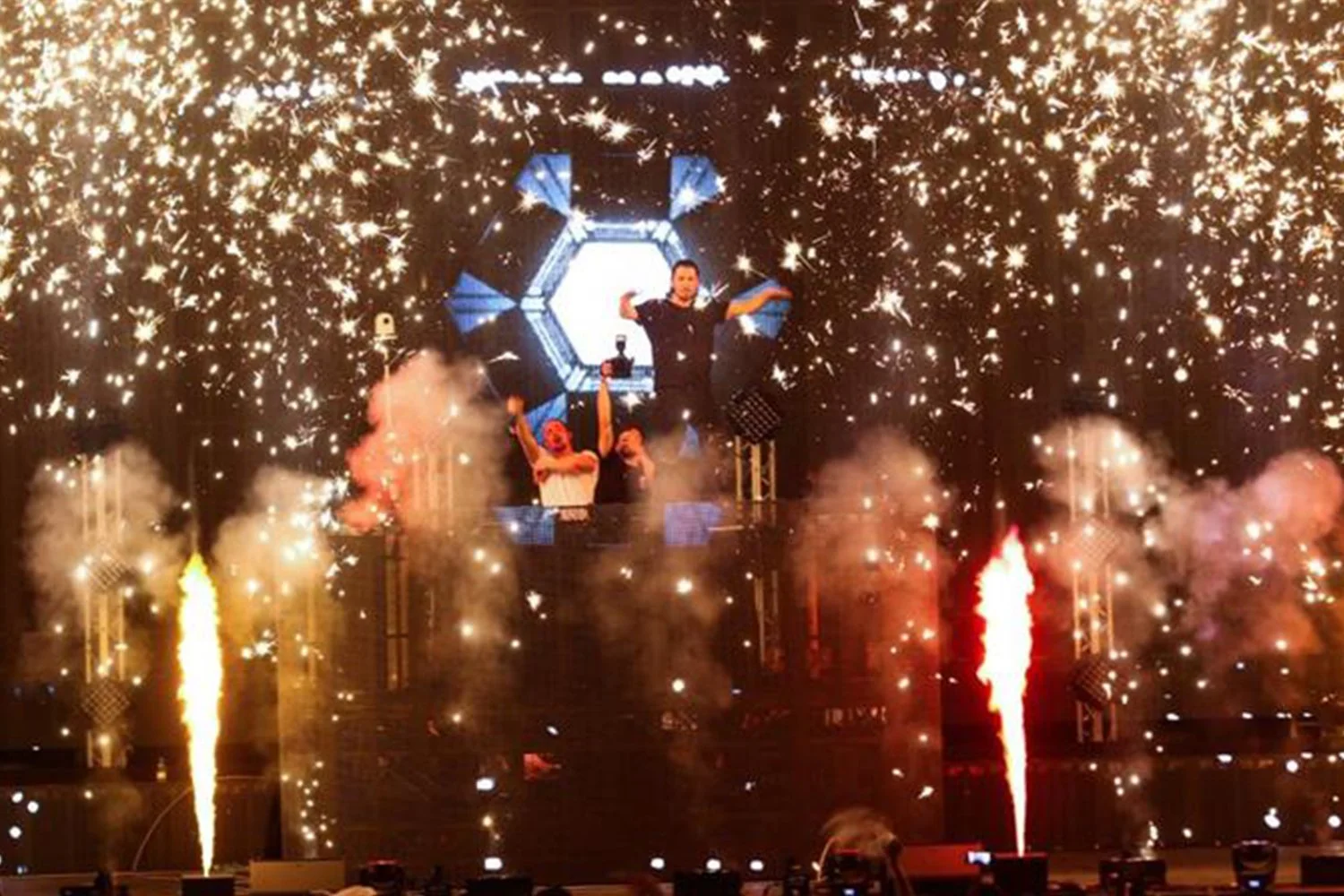 DJ performing at a concert with pyrotechnics and fireworks, large screen displaying a hexagon pattern in the background