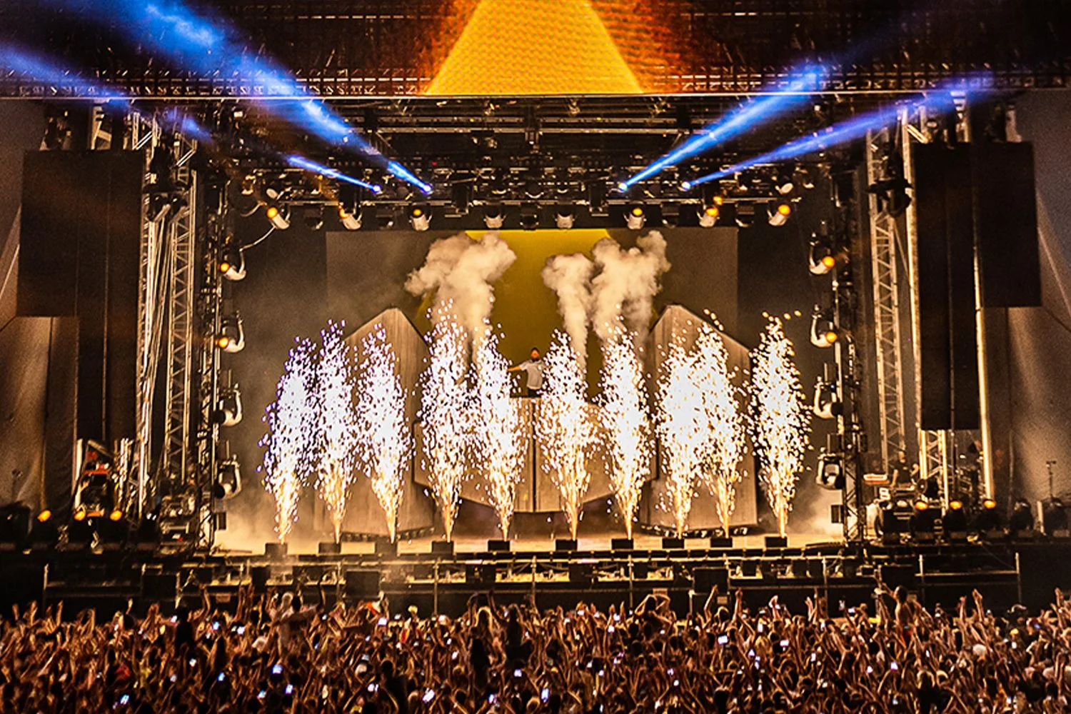 A large concert stage with fireworks, smoke, and bright lights, filled with a lively crowd taking photos.