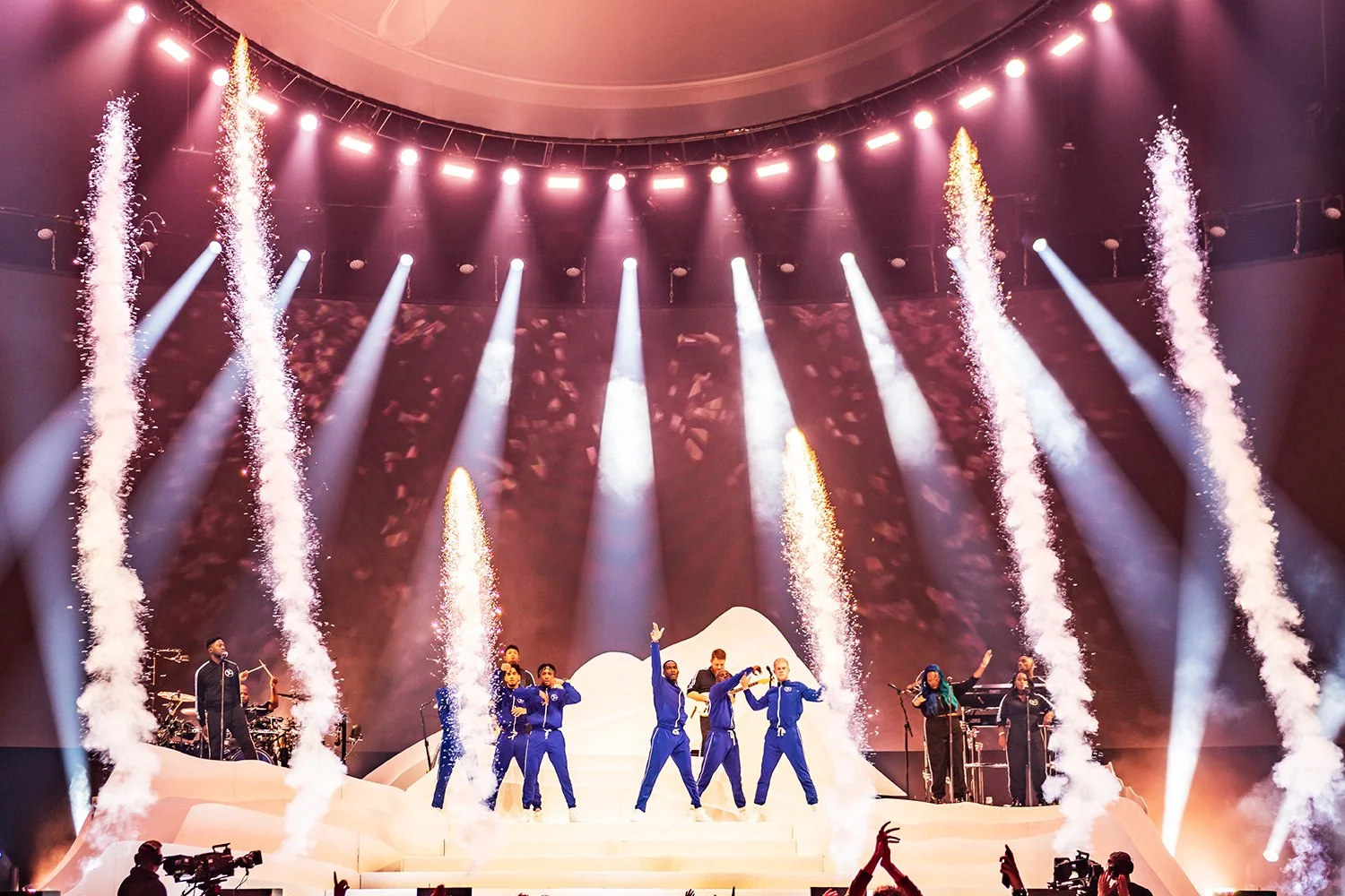 A concert stage with performers dressed in blue standing on white steps, with fireworks and colorful lights creating a dramatic atmosphere.