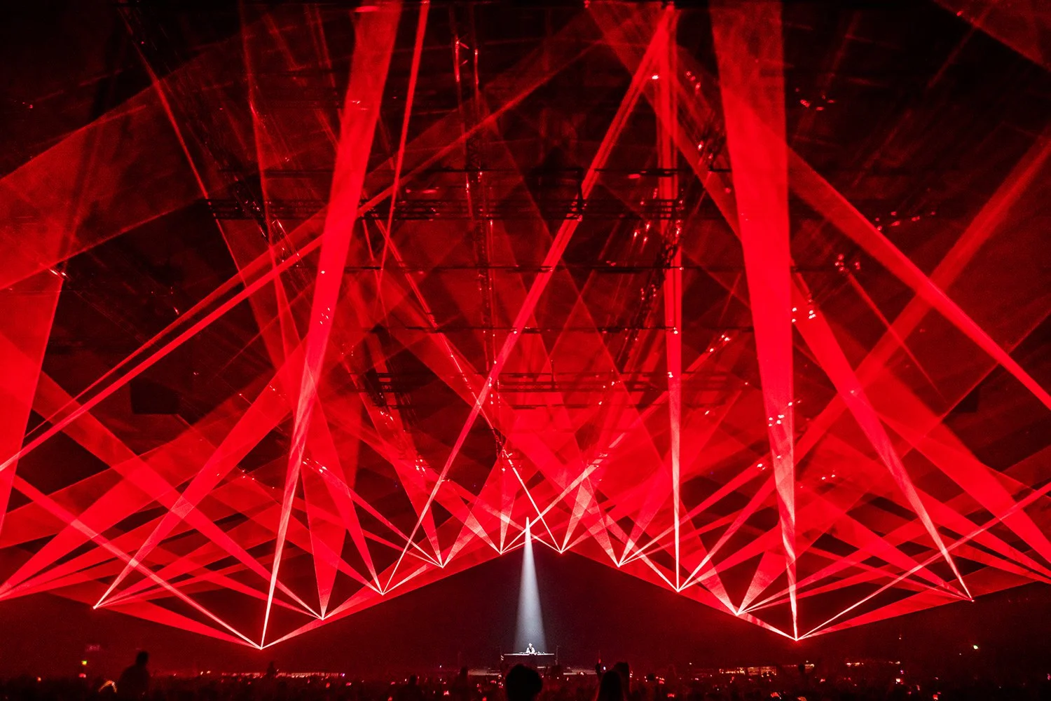 A concert scene with red laser lights creating intricate patterns above a DJ or performer on stage, with silhouettes of audience members in the foreground.