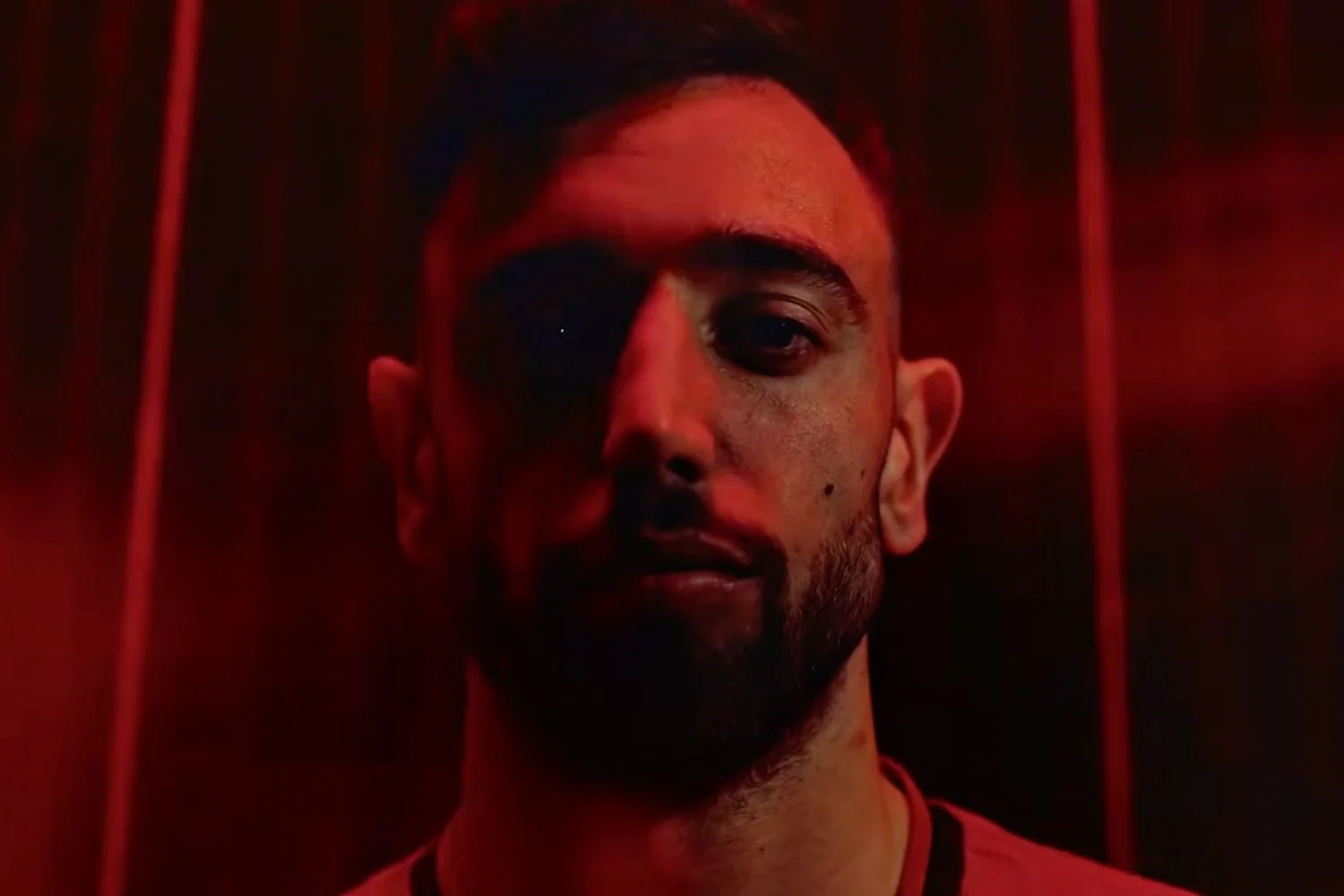 A man with an eye patch over his right eye, standing in front of a red background, with vertical red bars visible behind him.