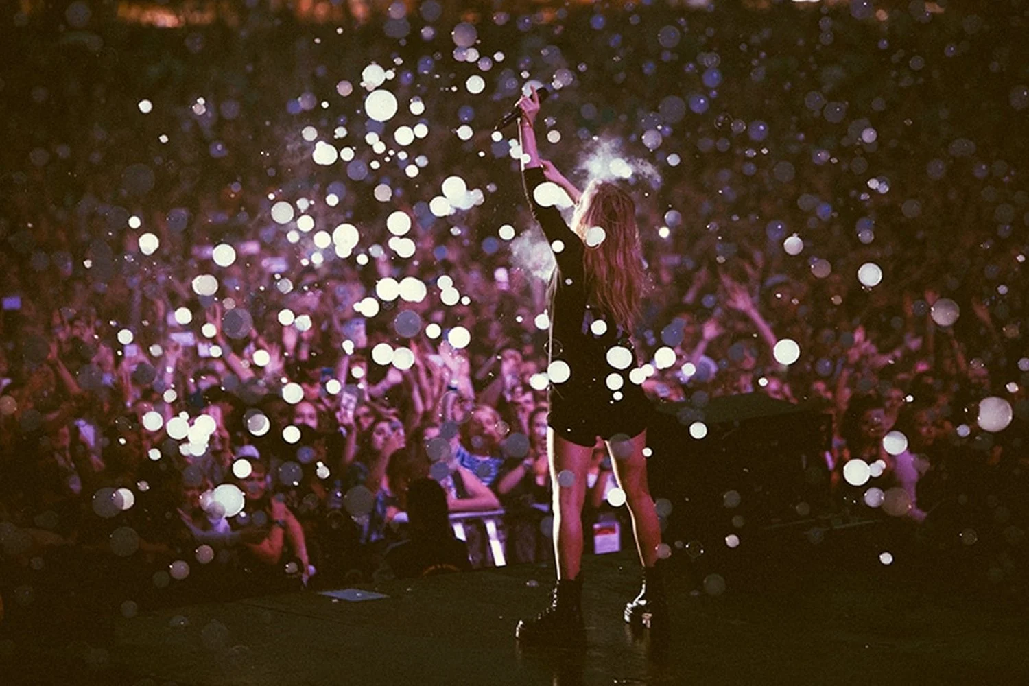 A female performer on stage at a concert, holding a microphone and raising her hand amidst a large crowd with floating light orbs and pinkish-purple lighting.