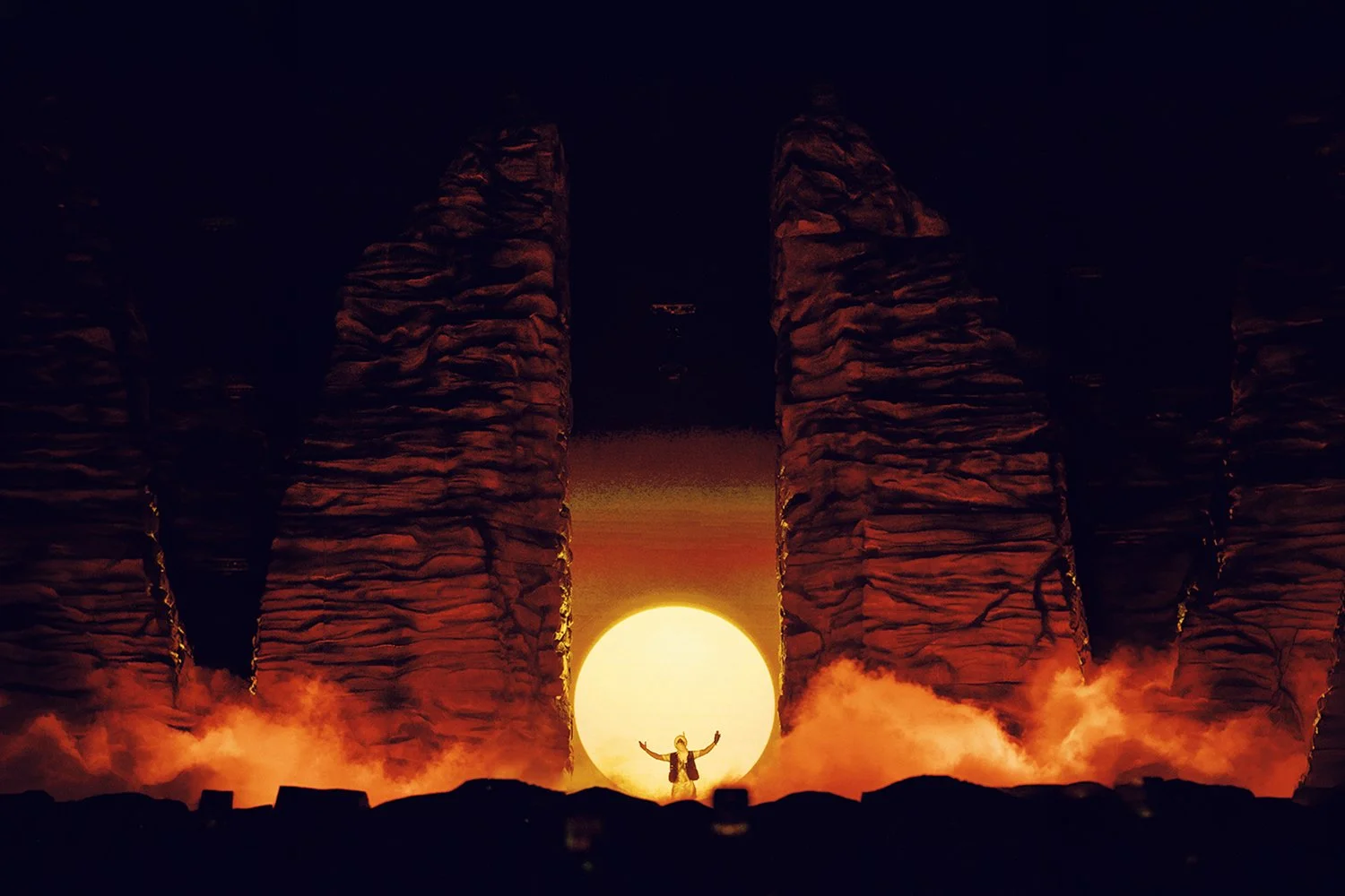 Person standing with arms raised at sunrise behind large rock formations with steam or smoke at their base.