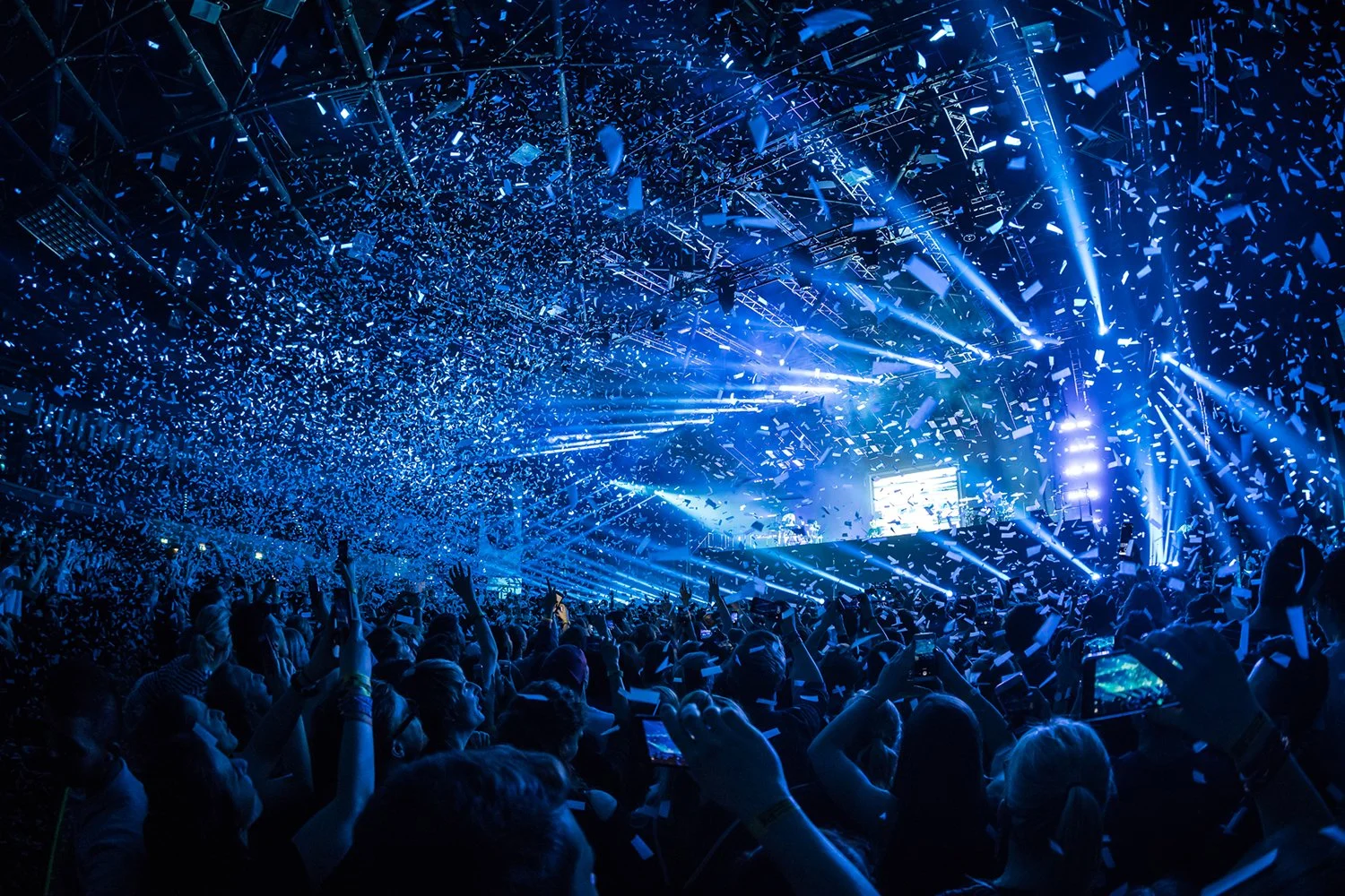 A concert with a large crowd, bright blue stage lights, confetti falling from the ceiling, and people holding up their phones.