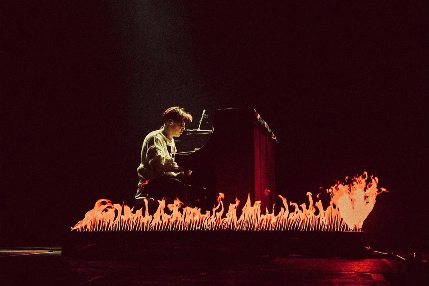 A person playing a black grand piano on a stage with flames surrounding the base of the piano, in a dark setting with minimal lighting.