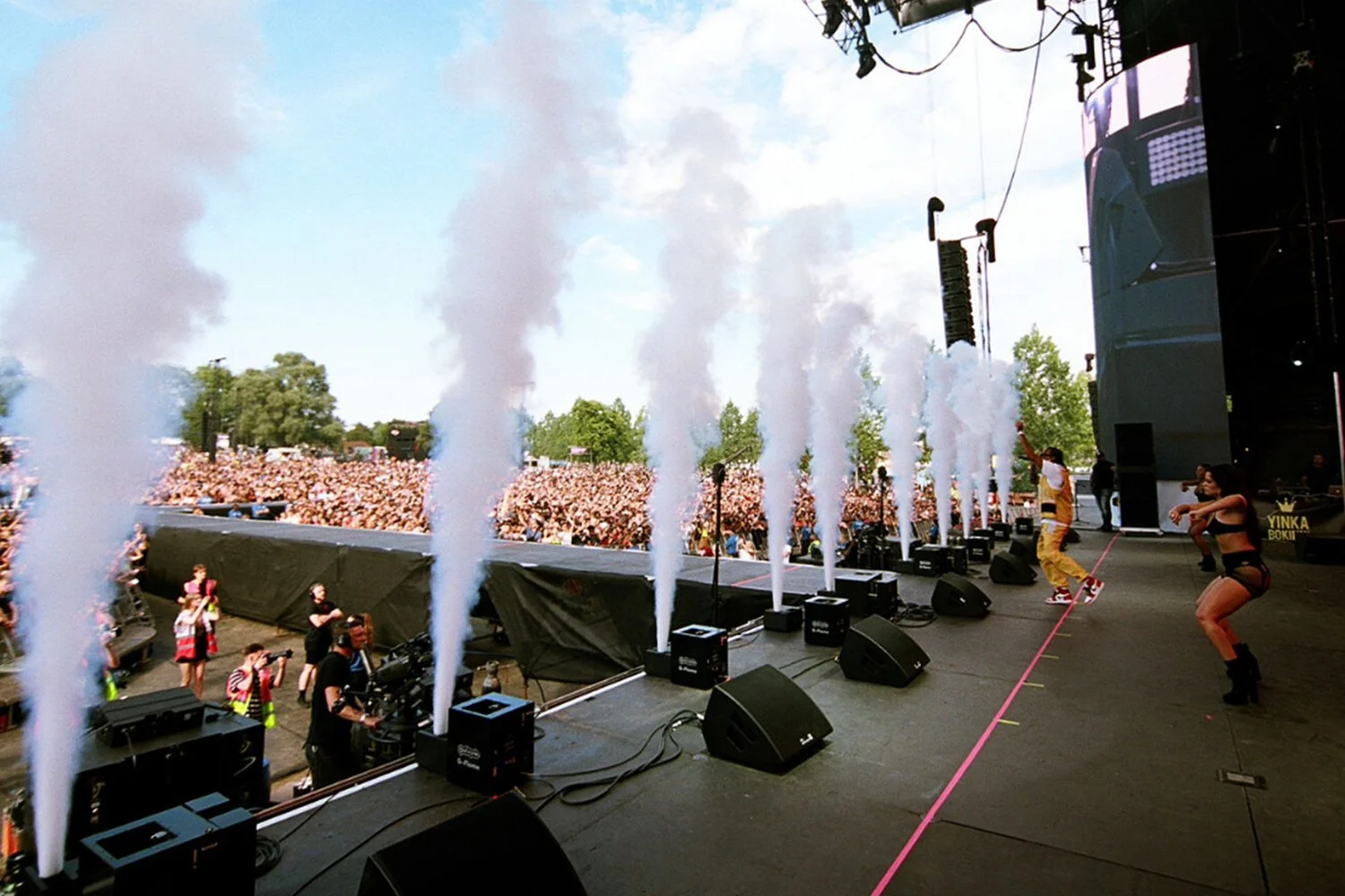 View of a large outdoor concert stage with smoke effects, performers dancing, and a giant crowd in the background on a sunny day.
