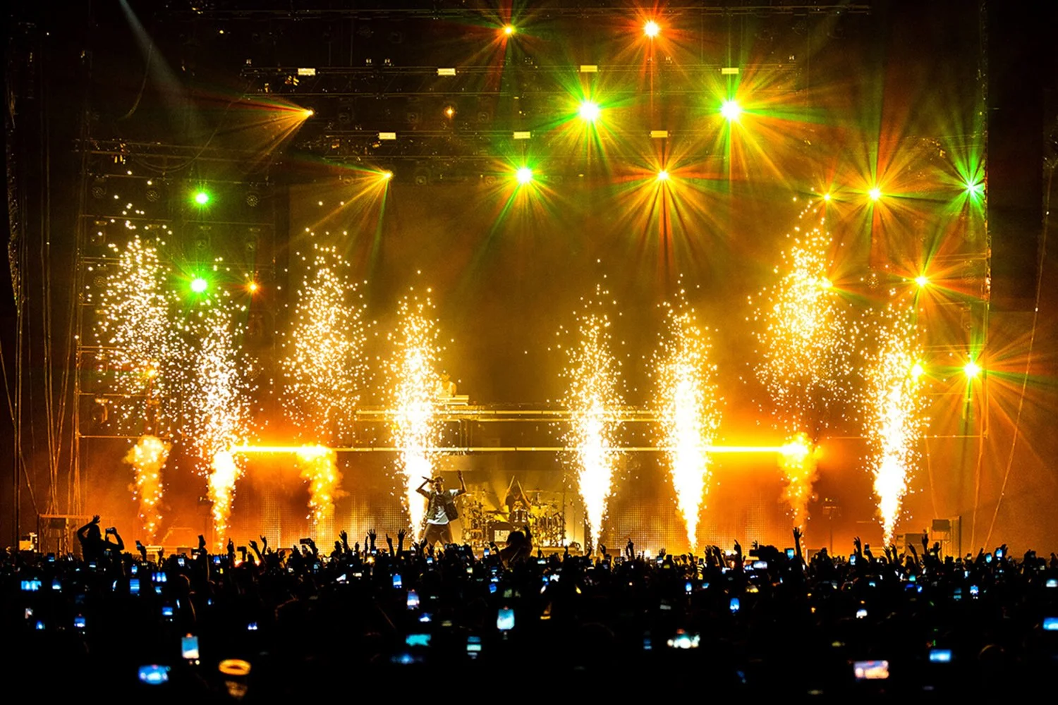 Live concert with bright yellow-green spotlights, pyrotechnics, and an audience with many people holding up phones