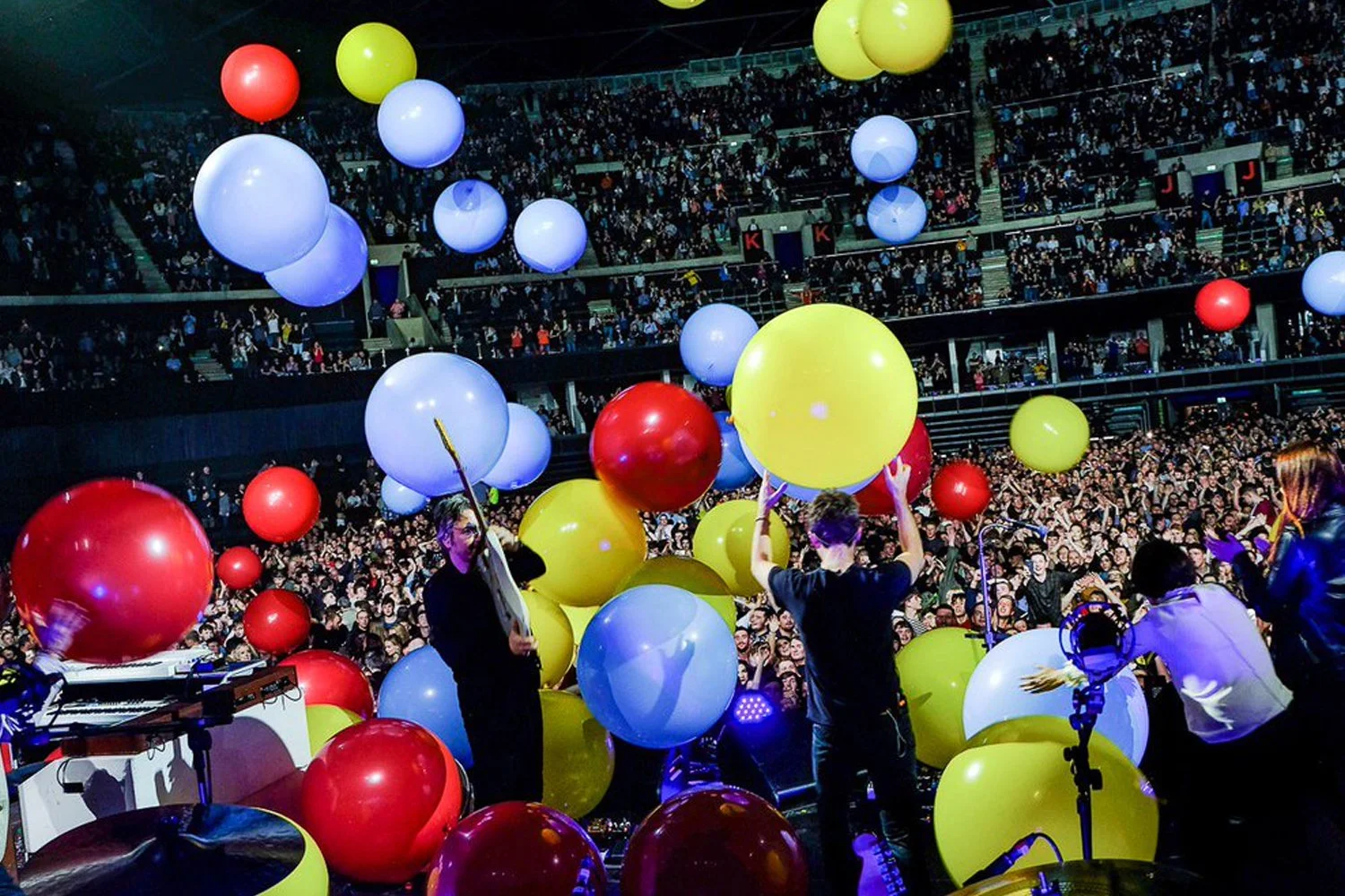 A concert stage with performers surrounded by large, colorful balloons in red, yellow, blue, and white, with a large crowd in the background.