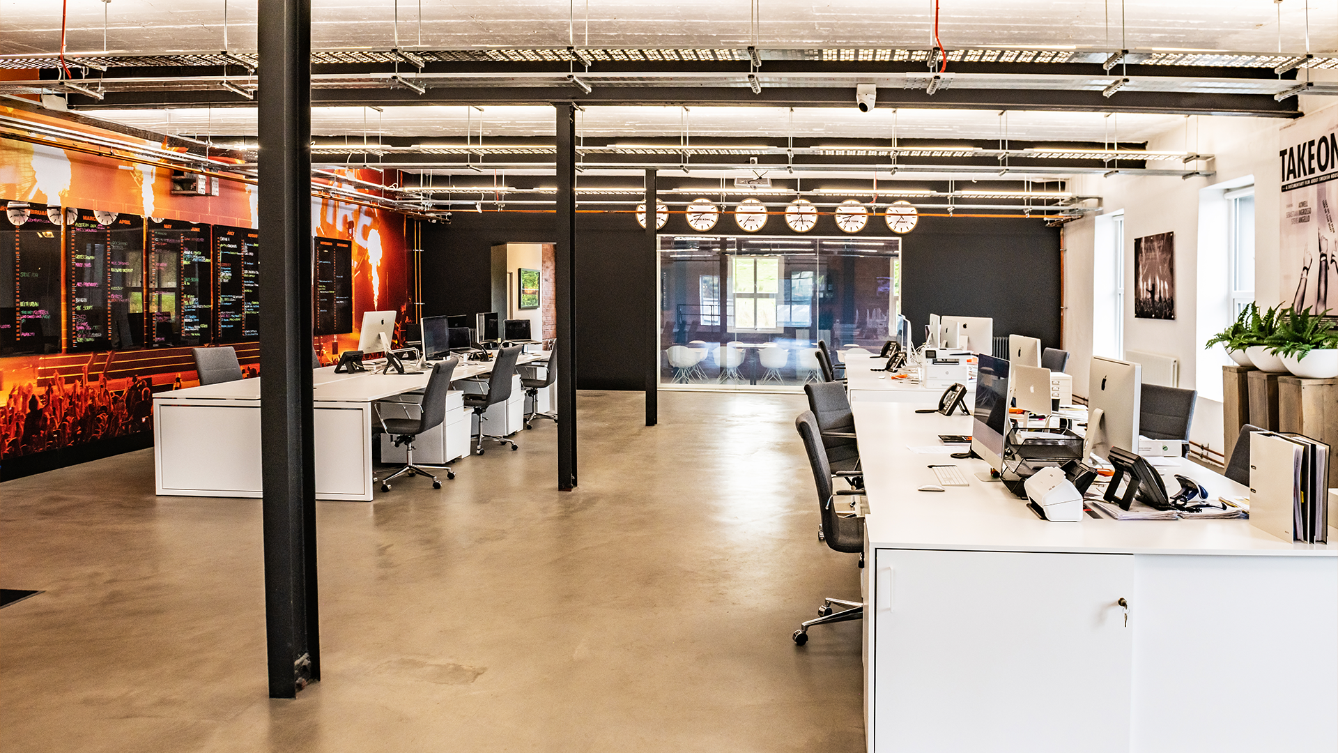 Modern open-plan office with white desks, black office chairs, desktop computers, and plants, divided by a black partial wall, with multiple wall clocks and a large window