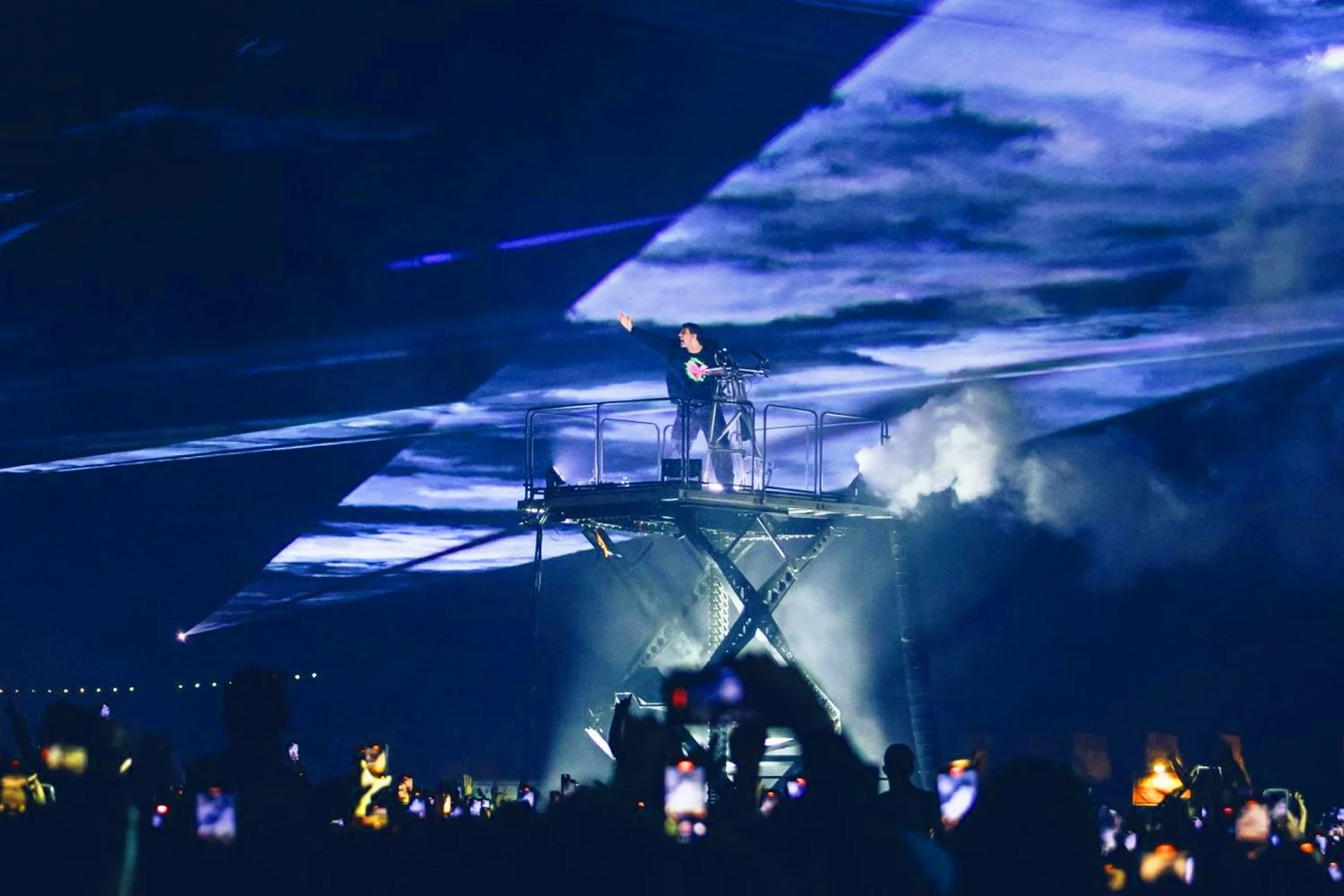 A performer on a raised platform during a concert, with audience members holding up phones, and dramatic lighting creating a vibrant atmosphere.
