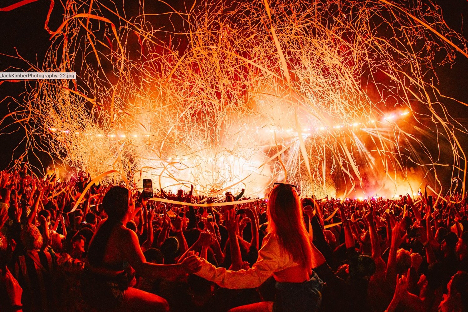A large crowd of people at a concert or festival during nighttime, with fireworks or pyrotechnics lighting up the sky and streamers or confetti falling over the audience.