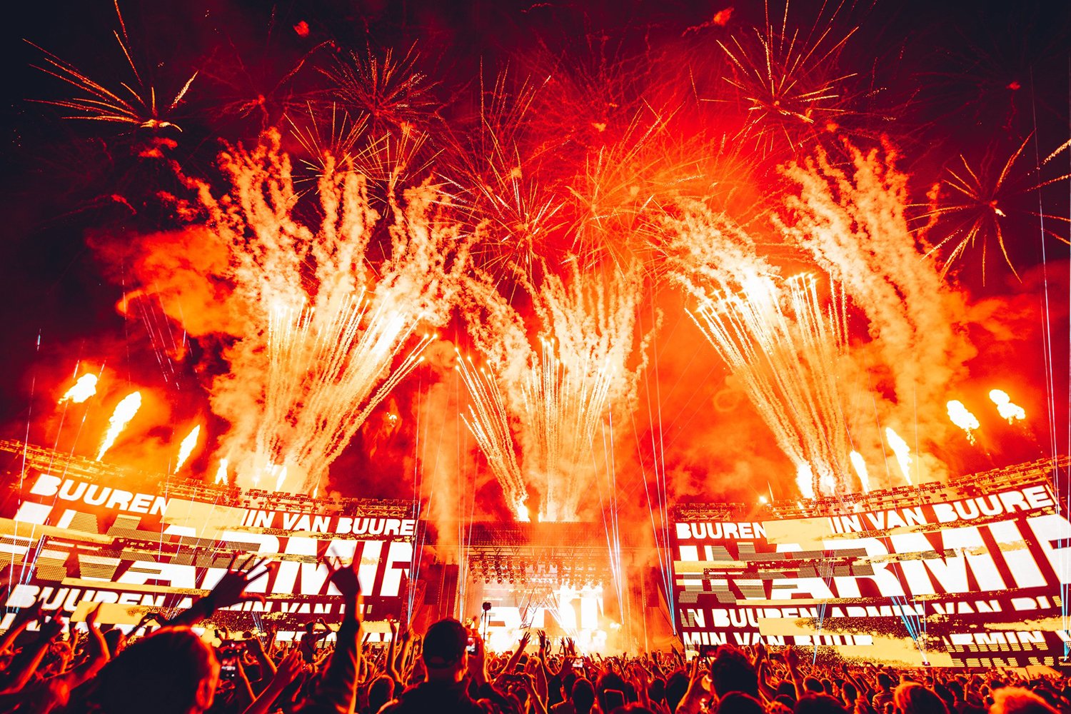 A large crowd watching a fireworks display at a concert or festival, with bright orange and red fireworks lighting up the night sky and screens showing text in Dutch.