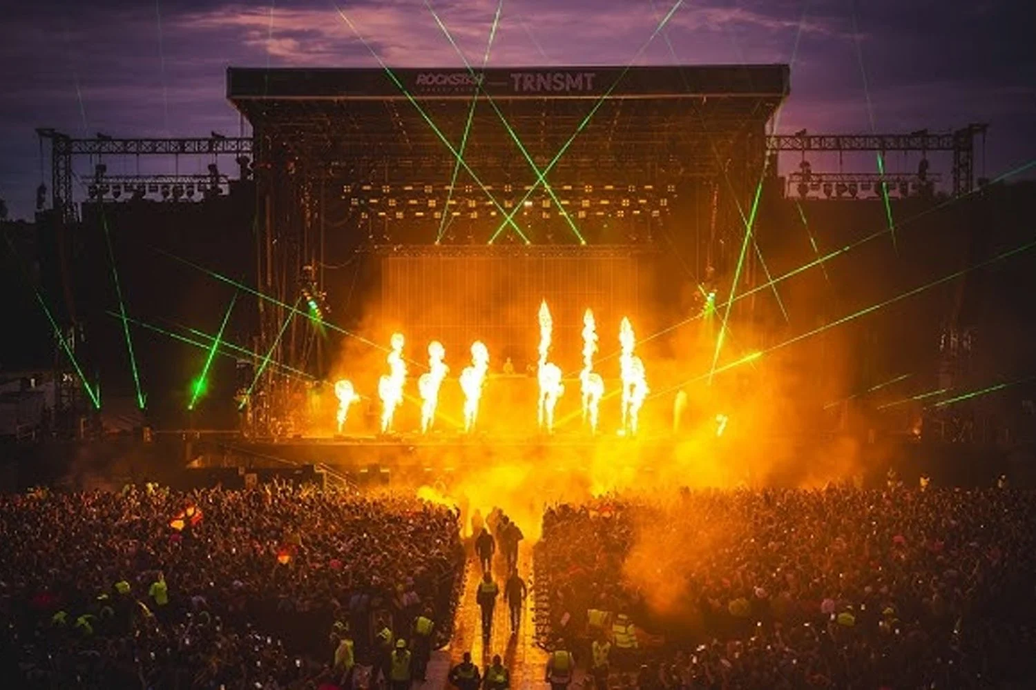 A large outdoor concert stage with fireworks and laser lights, surrounded by a massive crowd of spectators during dusk.