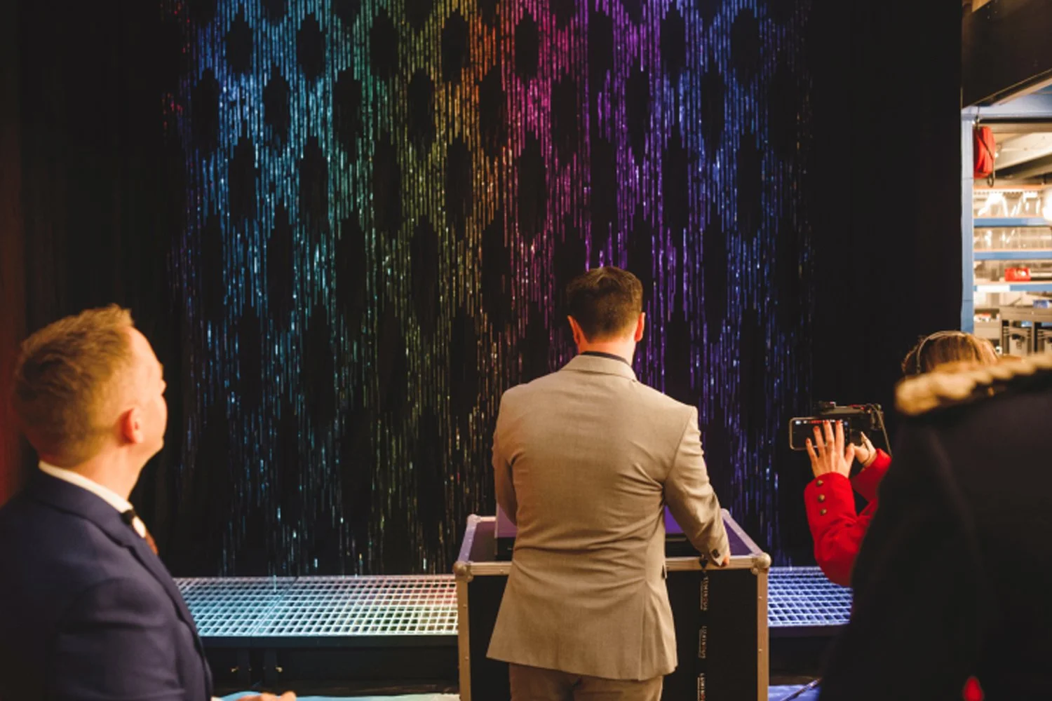 People observing a man in a light gray suit operating a device in front of a black curtain with colorful, shimmering vertical lines.