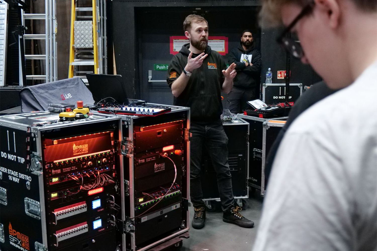 A man with a beard and a black hoodie explaining something to a person in a white shirt in a production or technical environment with computer equipment, racks, and tools around.