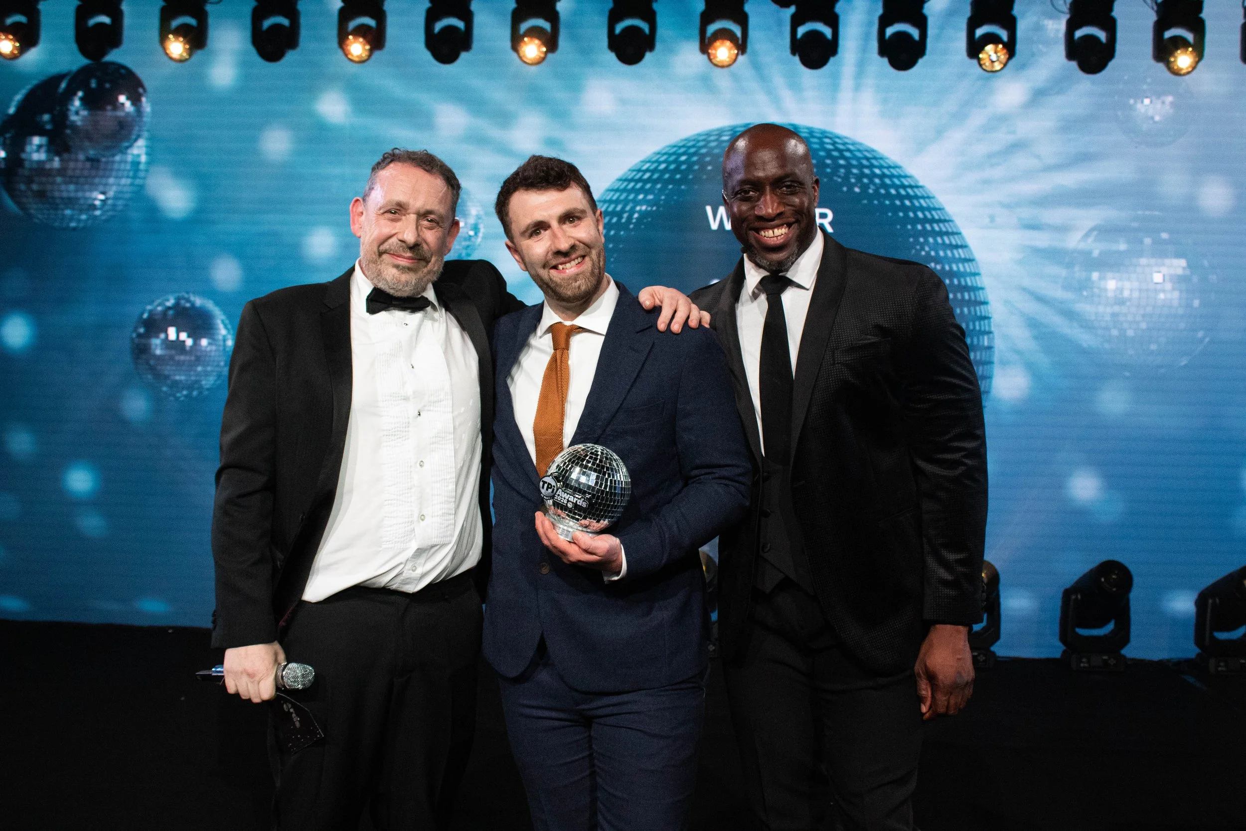 Three men dressed in formal attire, two wearing tuxedos and one in a suit, standing on stage at an awards ceremony. The man in the middle is holding a glass award, and there is a disco ball on the background with stage lights overhead.
