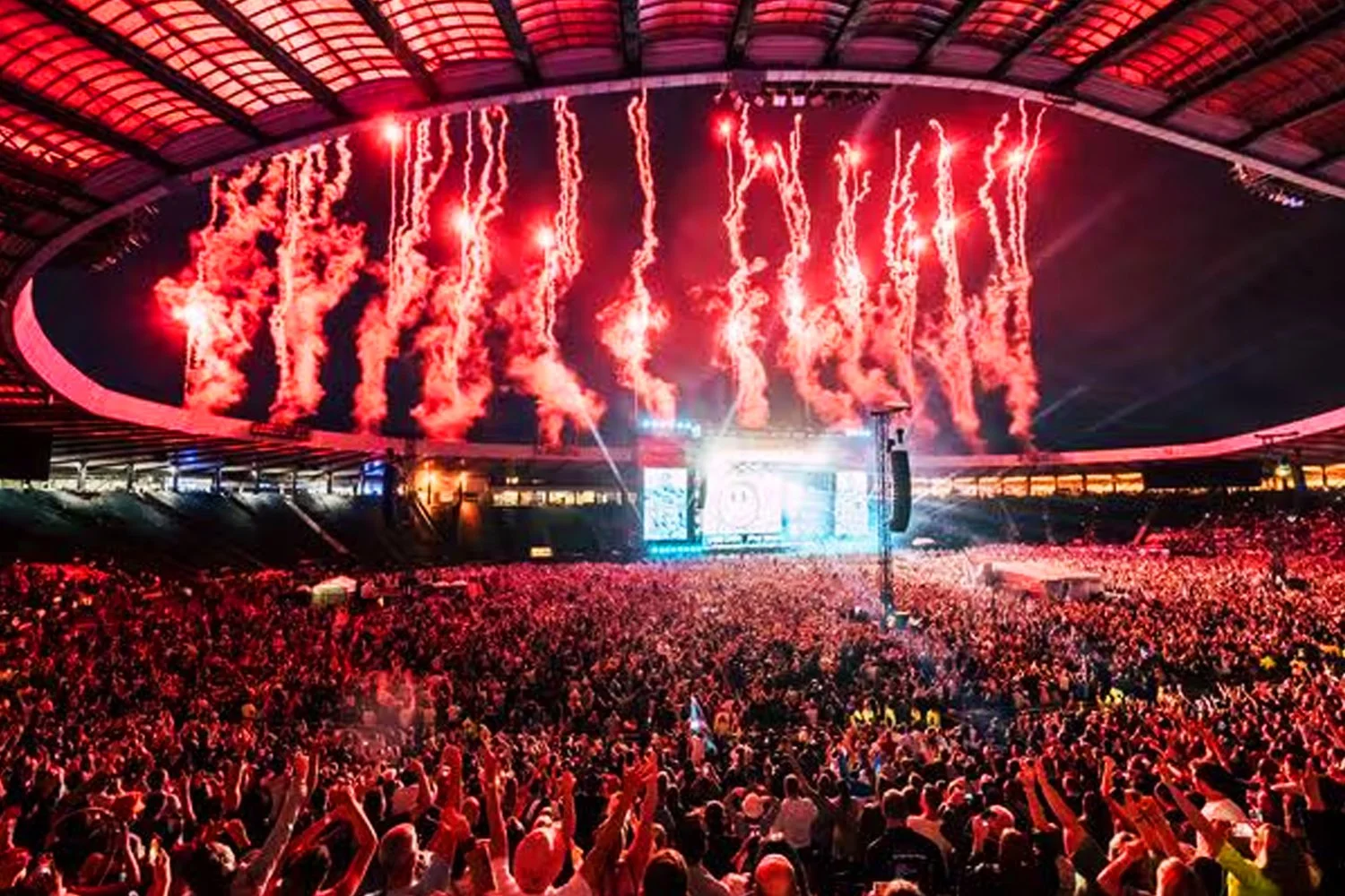 A large crowd at a concert stadium or arena during a fireworks display with red and orange fireworks in the night sky visible from the stage area.