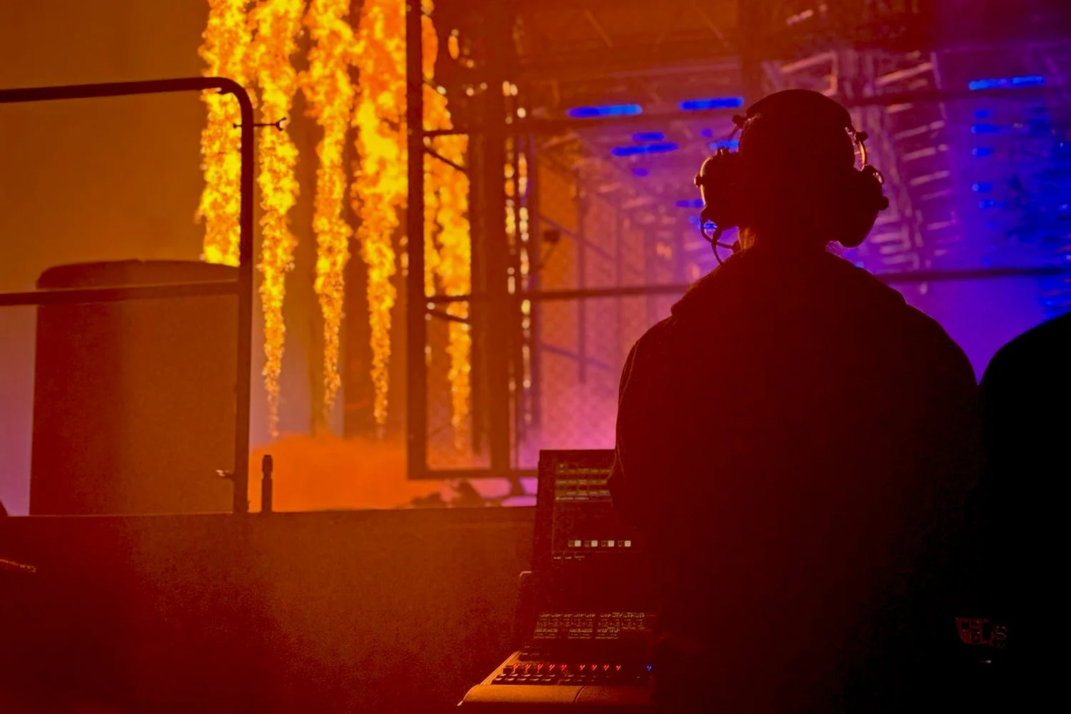 Silhouette of a DJ wearing headphones at a music event with vibrant orange, purple, and blue lighting effects and a fiery display in the background.