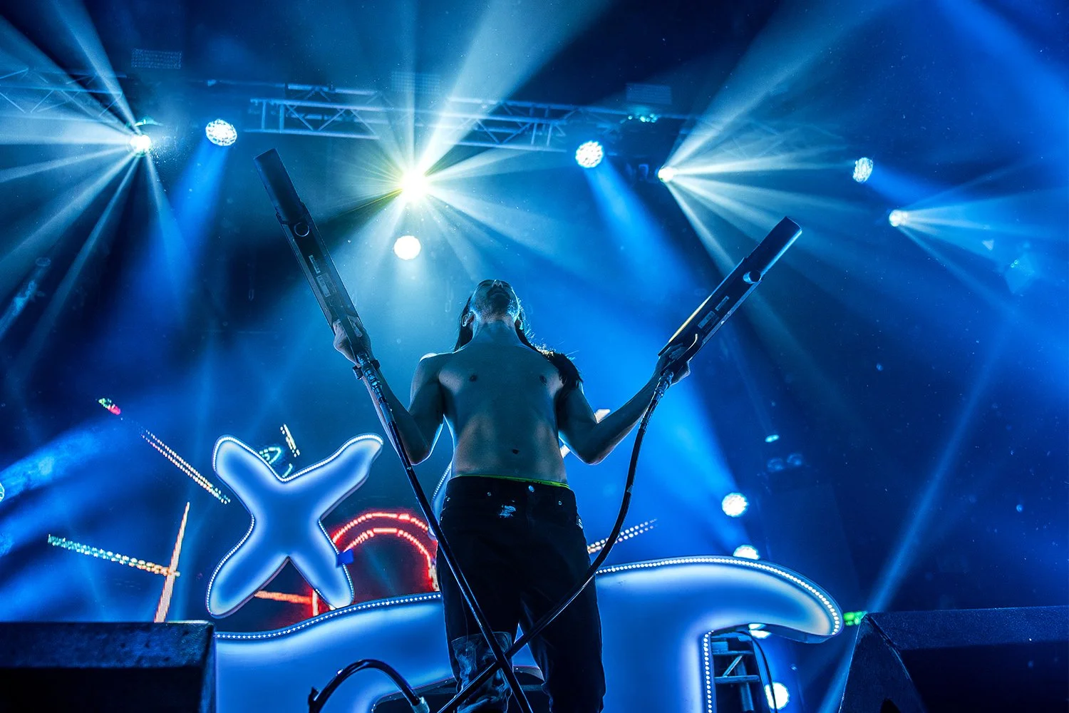 A shirtless DJ performing on stage with blue lighting, holding two large controller pads, surrounded by bright stage lights and a neon sign in the background.