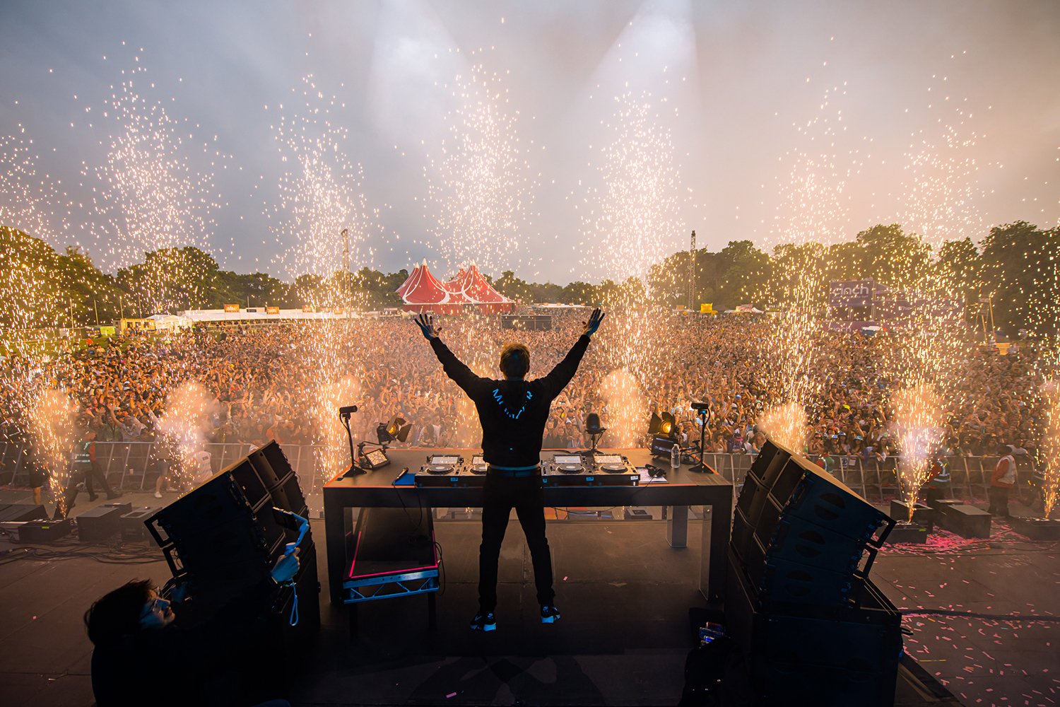 DJ performing on stage with arms raised at a concert, surrounded by pyrotechnics, with a large crowd in the background and trees beyond, during sunset.