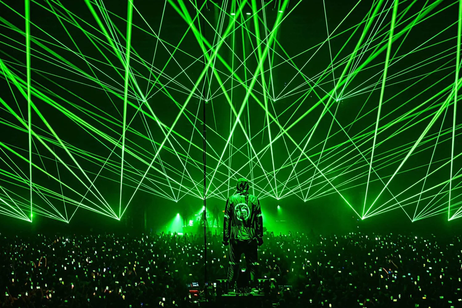 A live concert scene with a person on stage facing an audience under a display of green laser lights creating geometric patterns.