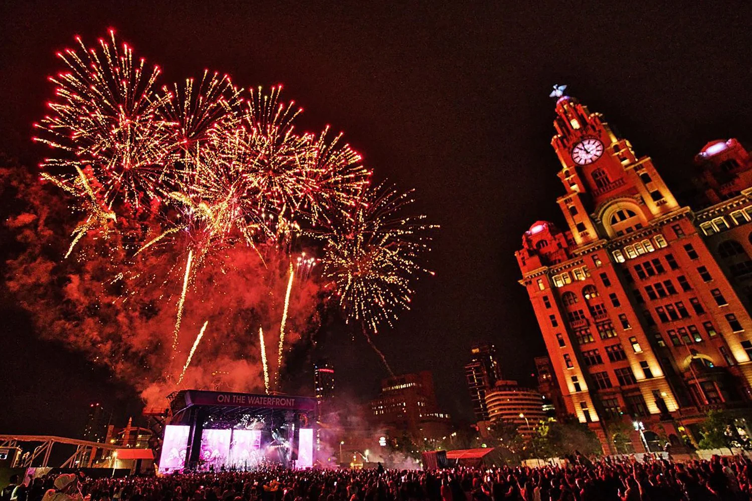 Fireworks display over a crowd during a celebration at night, with a historic building illuminated in red.