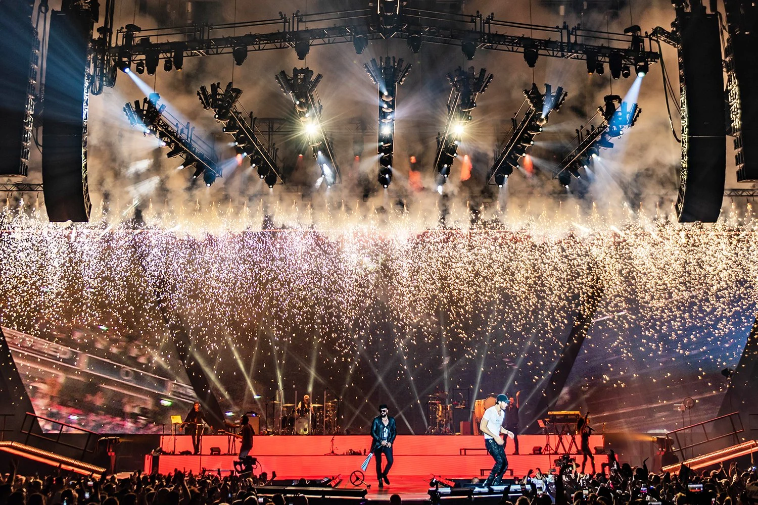 A concert stage with musicians performing, bright stage lights, pyrotechnics, and thick smoke, in front of a large audience at night.