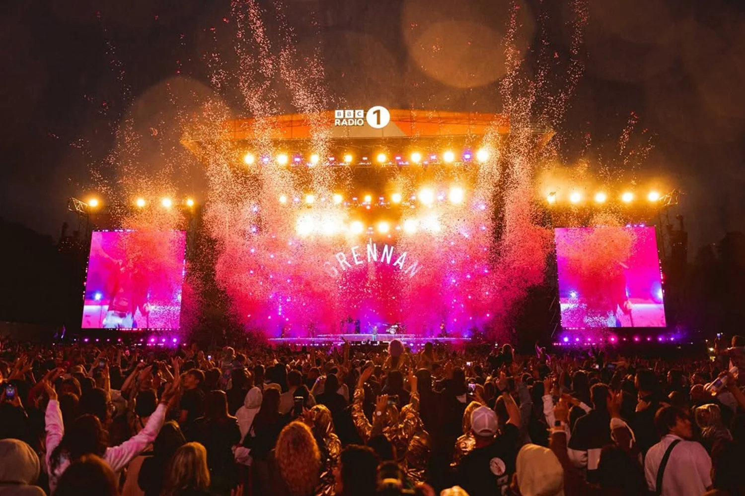 A large crowd at a concert or festival during nighttime, with a brightly lit stage displaying the name 'DRENNAN,' colorful lights, sparks and confetti in the air, and two large screens showing the audience.