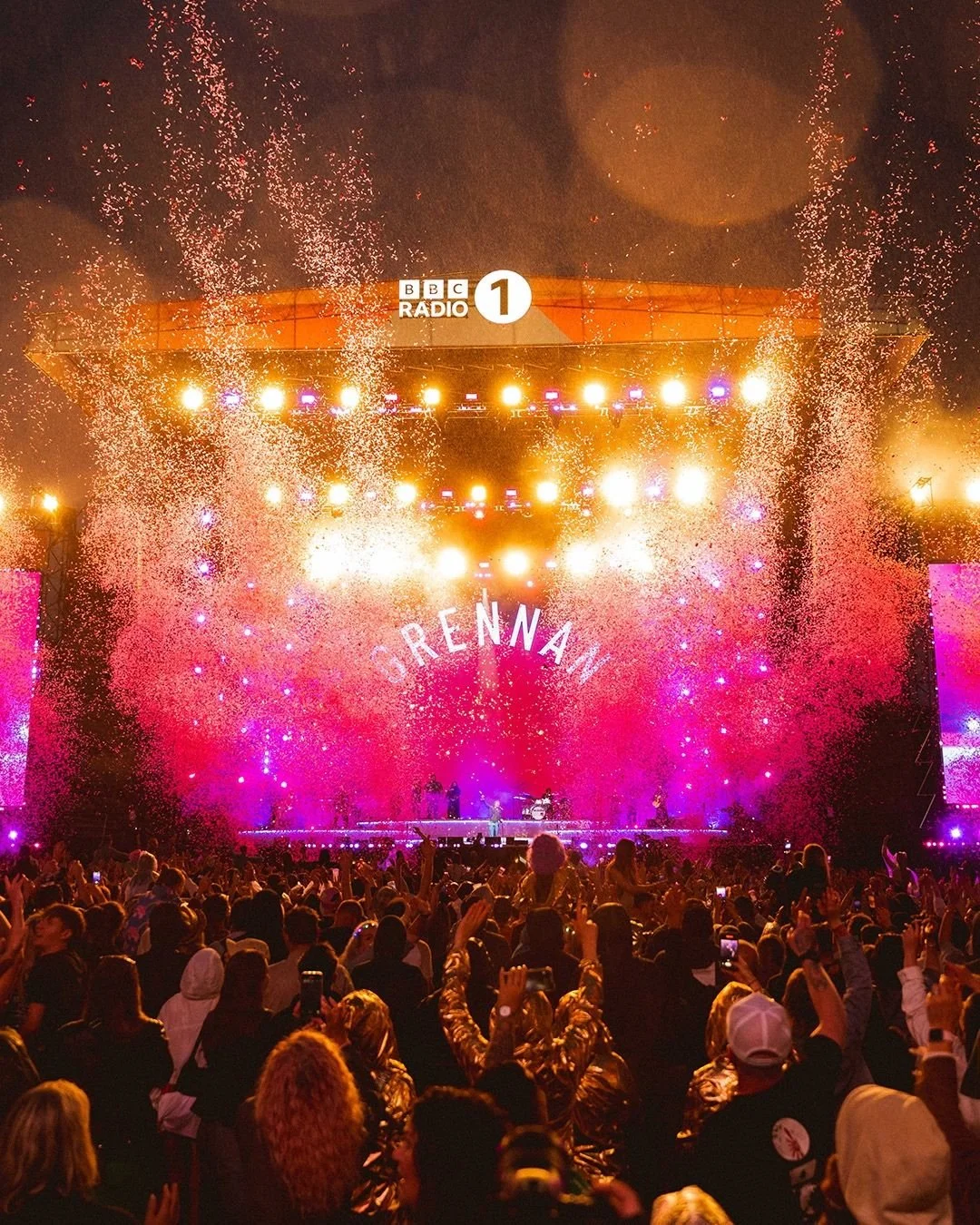Crowd at an outdoor concert with fireworks, stage illuminated in pink and purple, and the word 'DRENNA' displayed on the stage.