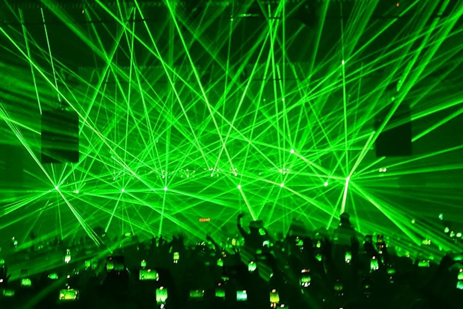 Green laser lights crisscross over a crowd at a concert or event, with many people taking photos or videos on their phones.
