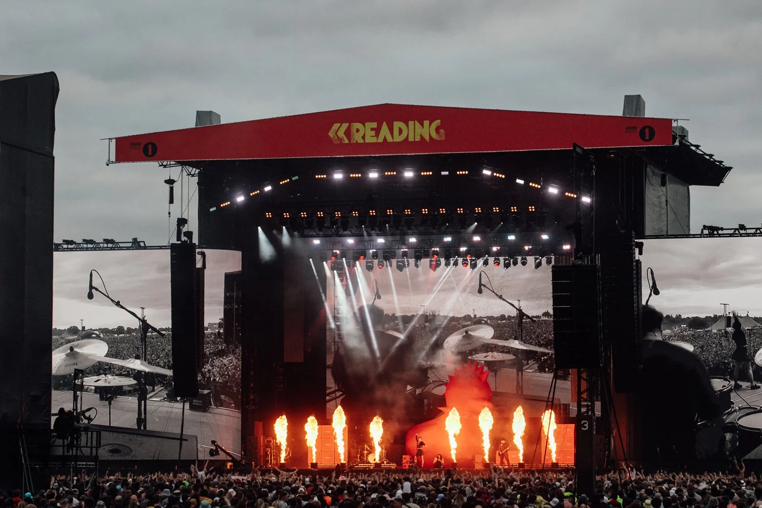 A large outdoor music concert with a stage illuminated by bright lights and pyrotechnics, and many people in the crowd watching.