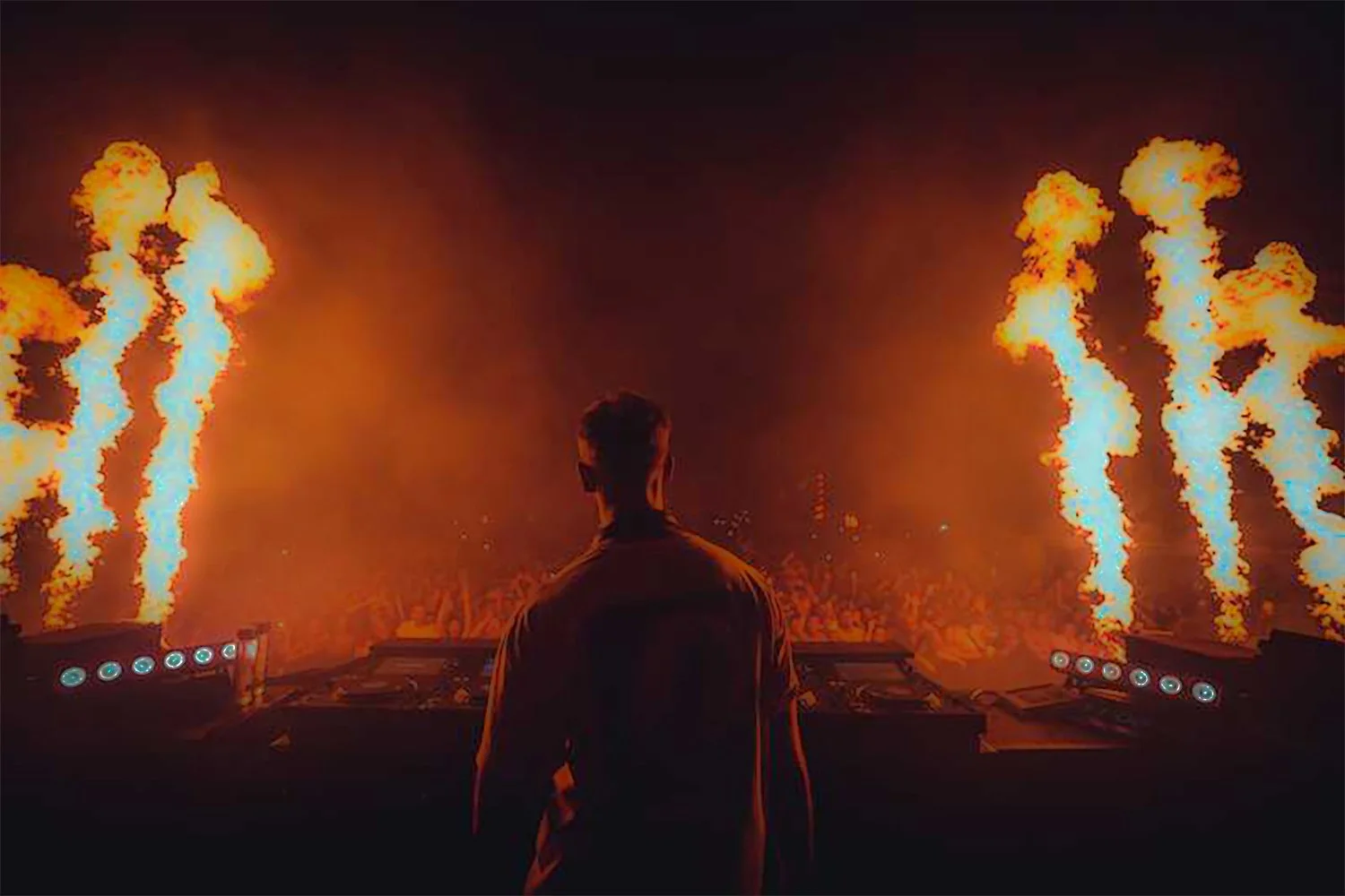 A DJ stands in front of a crowd with large flames shooting into the air on each side during a night concert.