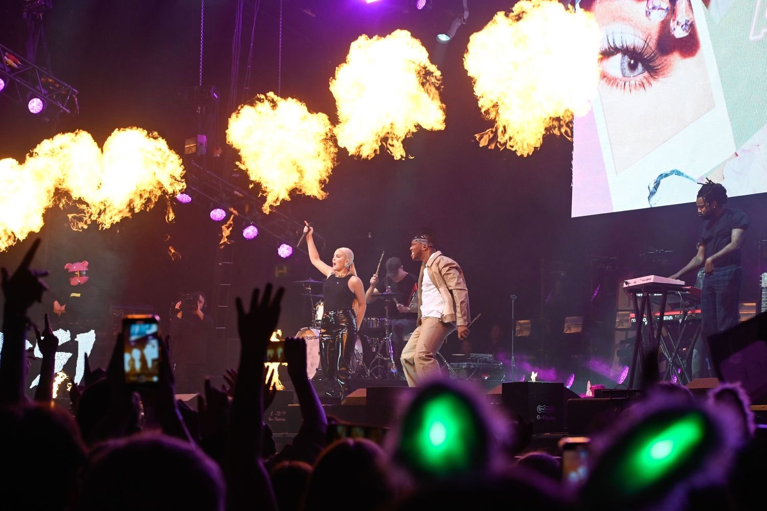 Music performance on stage with fire effects, featuring a woman in black leather pants, a man in beige pants, a drummer, and a keyboardist.