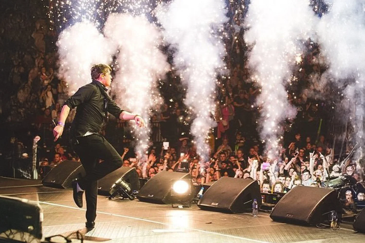 A performer on stage during a concert, holding a microphone, with colorful fireworks or pyrotechnics behind them and a crowd of fans cheering in the background.
