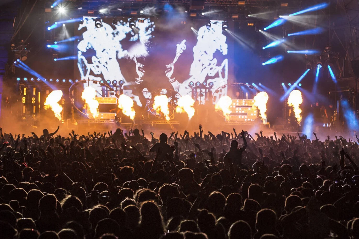Crowd at a concert with bright stage lights, pyrotechnics, and a large LED screen displaying abstract visuals.
