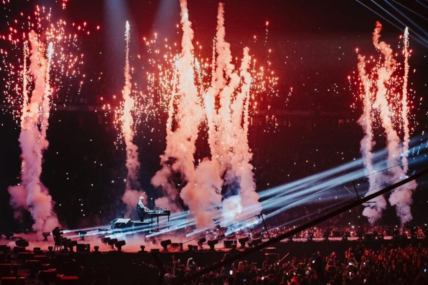 A concert stage with a DJ performing amidst red fireworks and smoke effects, bright spotlights shining, and an audience watching from below.