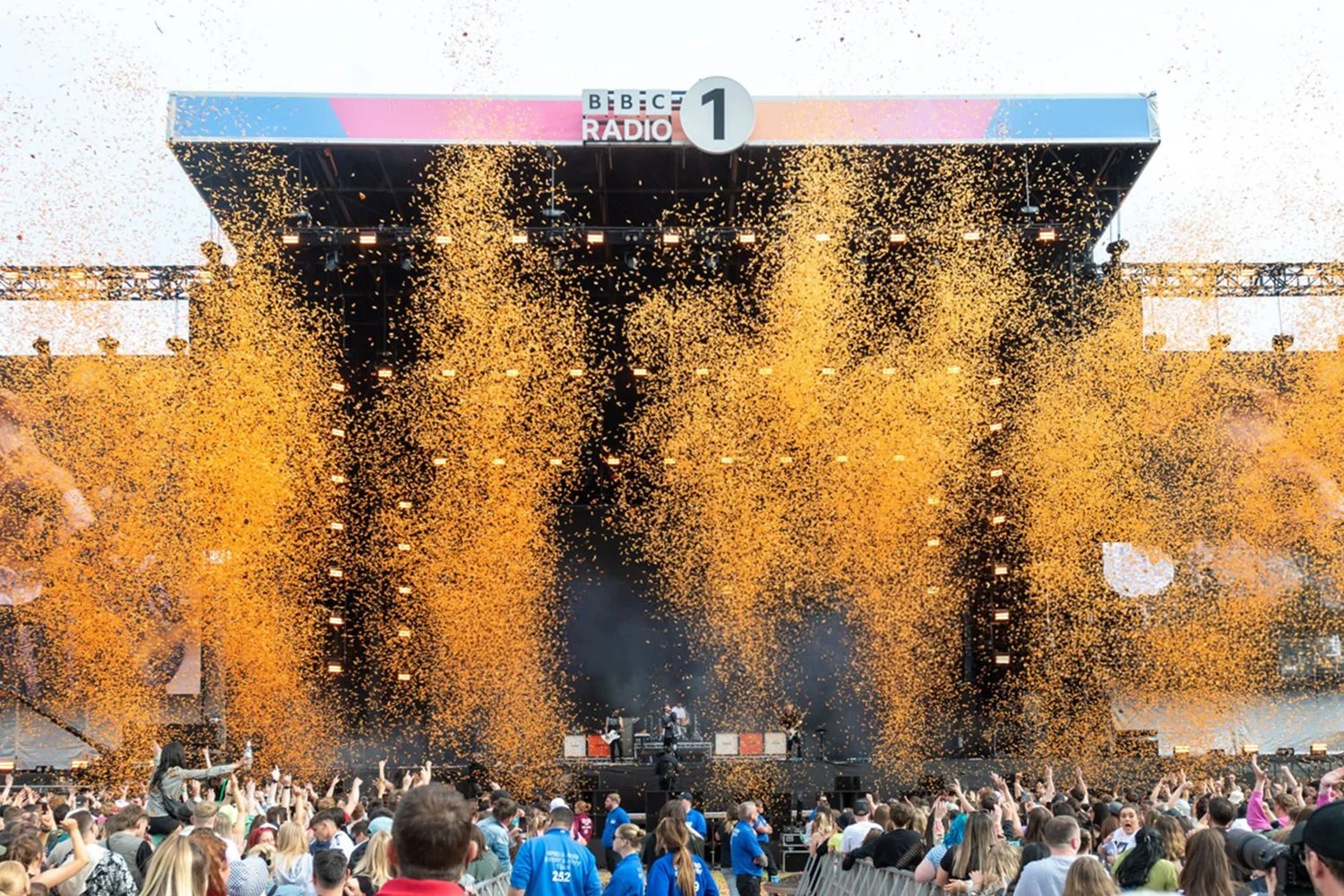 Crowd attending a music concert outdoors with a large stage, golden confetti falling, and a band performing.