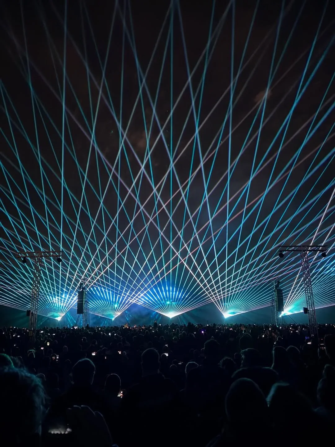 A concert or large event with a crowd watching a laser light show in the night sky, with blue and white laser beams crossing above the stage.