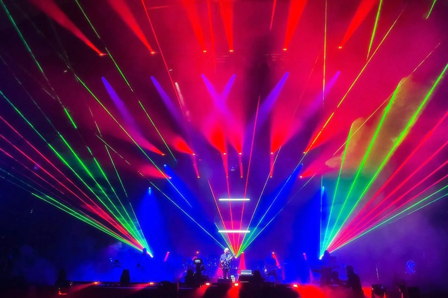 A concert stage with vibrant red, green, and blue laser lights creating dynamic patterns and a performer in the center with a guitar.