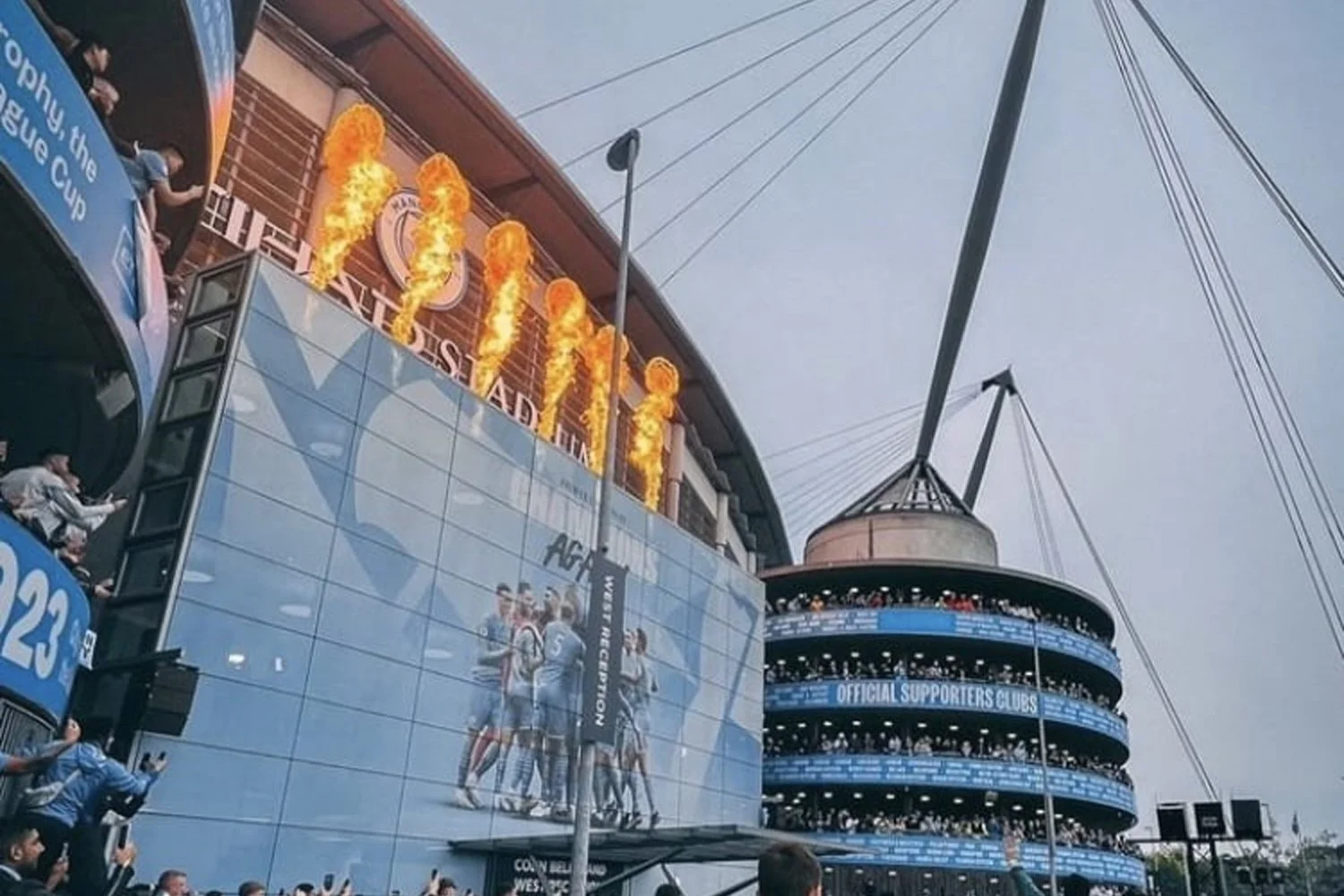 A large crowd at a stadium with a modern glass building and a circular structure. Flames shoot up from the building with an AC Milan logo, indicating a soccer match celebration.