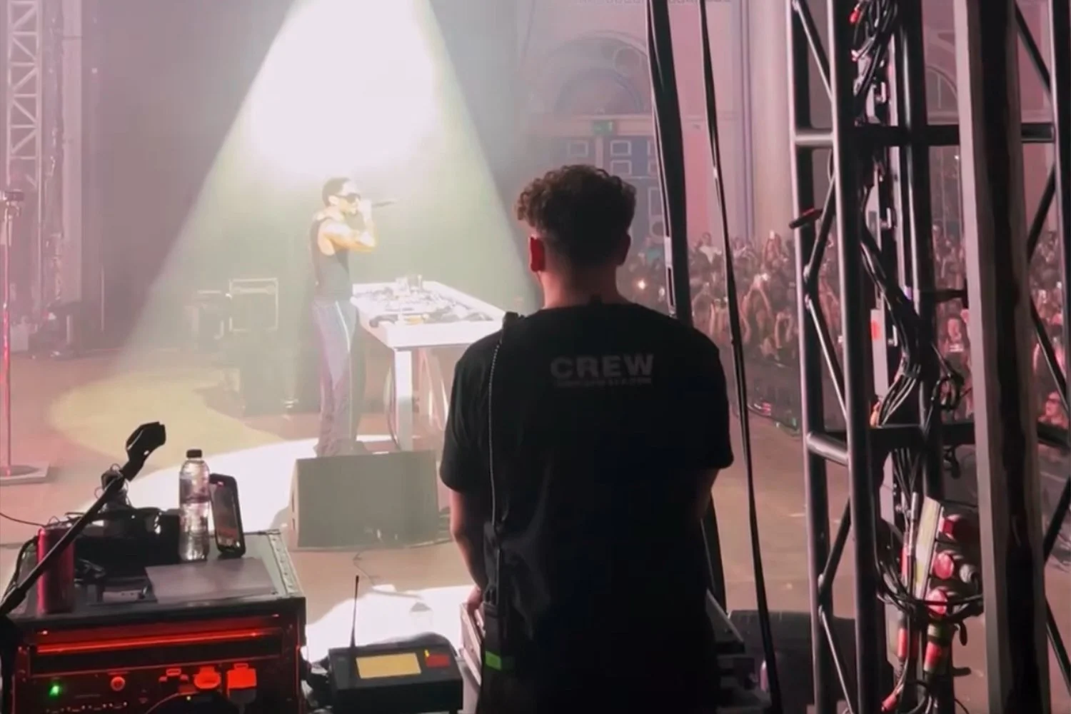 Behind the scenes view of a DJ performing on stage, with a person in the foreground wearing a 'CREW' shirt, technical equipment, and an audience in the background.
